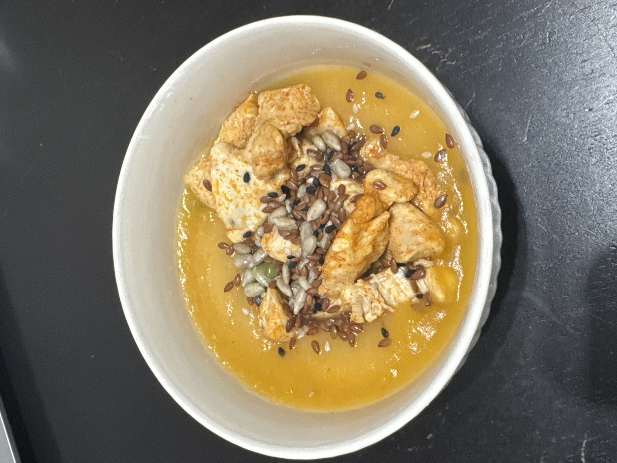 A top-down shot shows a bowl of yellow soup with a pile of chicken pieces and mixed seeds in the center. The bowl is white and sits on a black surface. No other subjects or objects are visible, and there is no indication of a specific location in Perm, Russia.