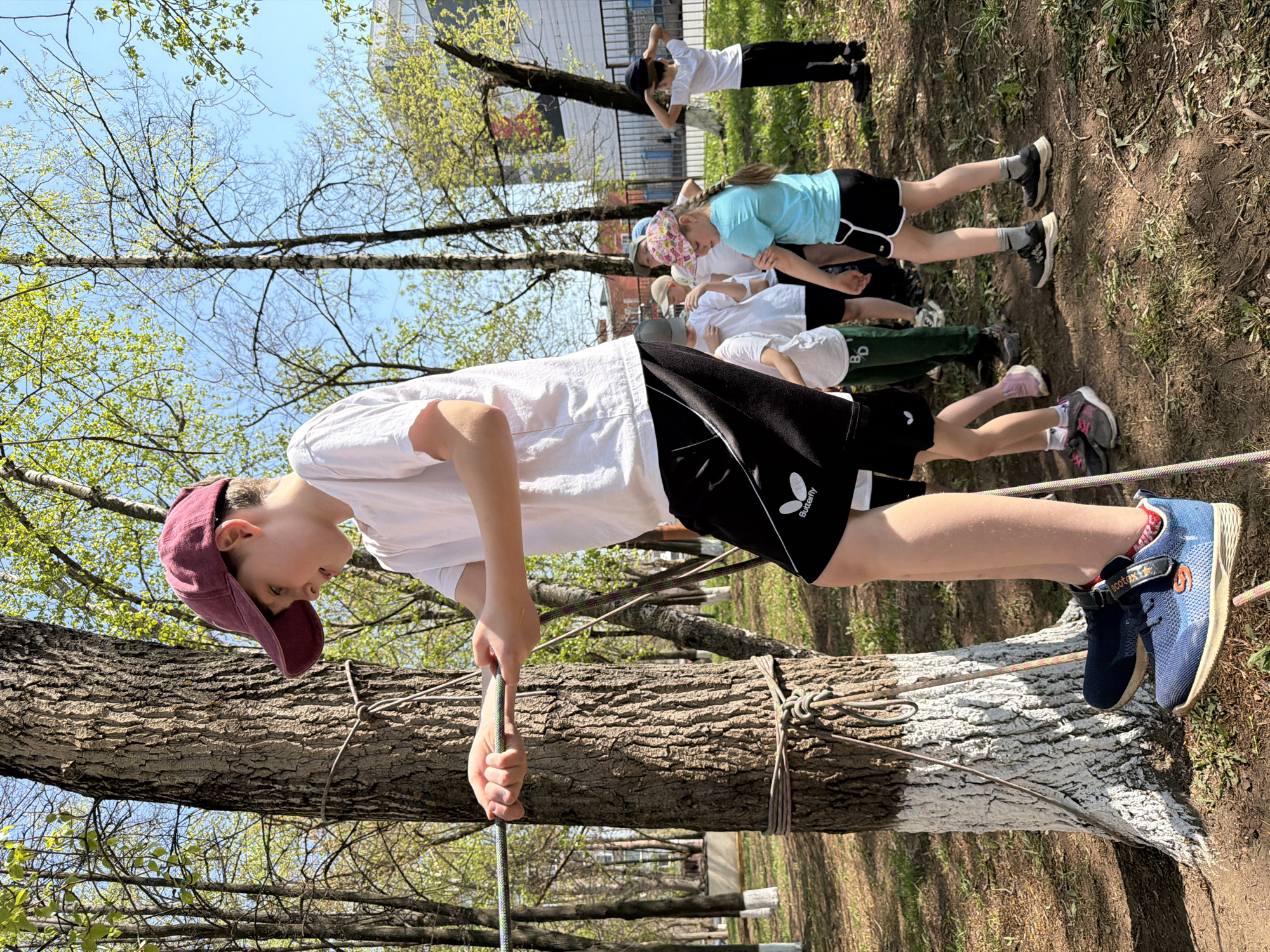 A daytime outdoor scene shows a group of children participating in what appears to be a rope course activity. A young person wearing a maroon baseball cap, a white t-shirt, and black shorts with a butterfly logo stands on a thick tree branch, gripping a rope. The branch is painted white on its lower portion and is part of an obstacle course that is tied to other trees. In the background, other children are lined up or engaged in the same activity, some wearing athletic shorts and t-shirts. The trees have new leaves budding, indicating springtime. The location is Izhevsk, Russia, based on the "BD" logo found on some clothing that are typically associated with a sports school in Izhevsk. A building is visible in the background behind the trees. The ground is covered with dirt and some sparse vegetation.