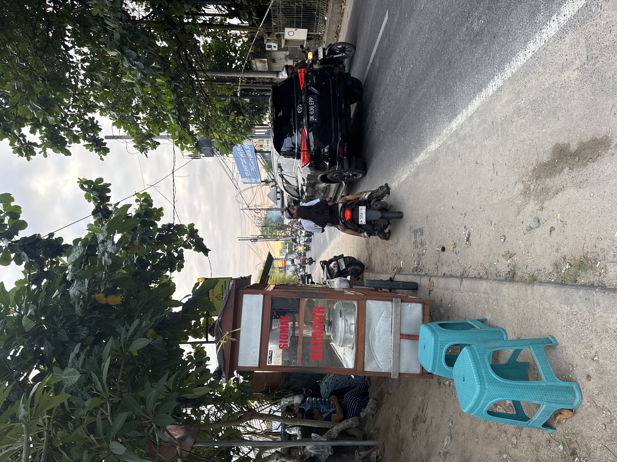 This media file captures a street scene in Kuta Selatan, Indonesia, likely during the daytime under an overcast sky. The foreground is dominated by a small food cart with a wooden frame and glass panels, displaying the text "SIOMAY BATAGOR" in red. Beneath the cart, several people are seated, appearing to rest or await service, with bottles of water visible. To the right of the cart are two turquoise plastic stools placed on the dusty ground.
Behind the food cart, the street is bustling with activity. A black car with the license plate "DK 636 EPP" is parked on the road, and a motorcycle is stopped nearby, with a person seated on it. Further down the street, a line of motorcycles and cars are visible, suggesting moderate traffic. A blue sign reads "SELAMAT DATANG DI PANTAI MUAYA JIMBAR", indicating the proximity to a beach. Trees with lush green foliage frame the left side of the image, partially obscuring the view of the sky and some overhead wires. The overall impression is of a typical street scene in a populated area, with food vendors and local transportation.