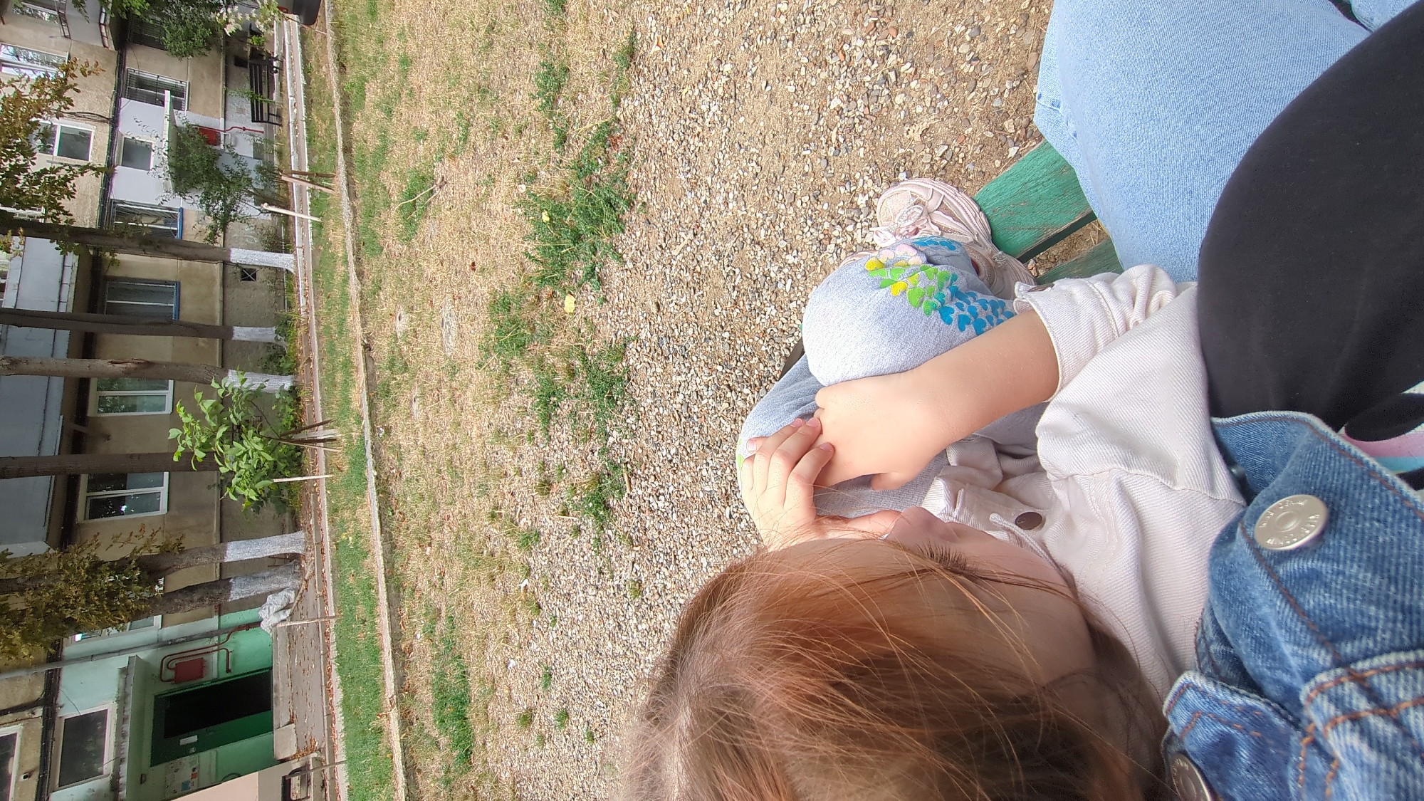 In Chișinău, Moldova, an outdoor scene unfolds with a child, seen from behind and slightly from the side, sitting on a green park bench. The child has fair hair and is wearing a light-colored jacket and light blue jeans adorned with a floral pattern. Their hands are clasped together in their lap, and they appear to be wearing earbuds. The background features a residential building with multiple windows and balconies, trees with white-painted bases, and a gravelly courtyard with patches of dry grass. A dark object, possibly another person's leg, is visible in the foreground.