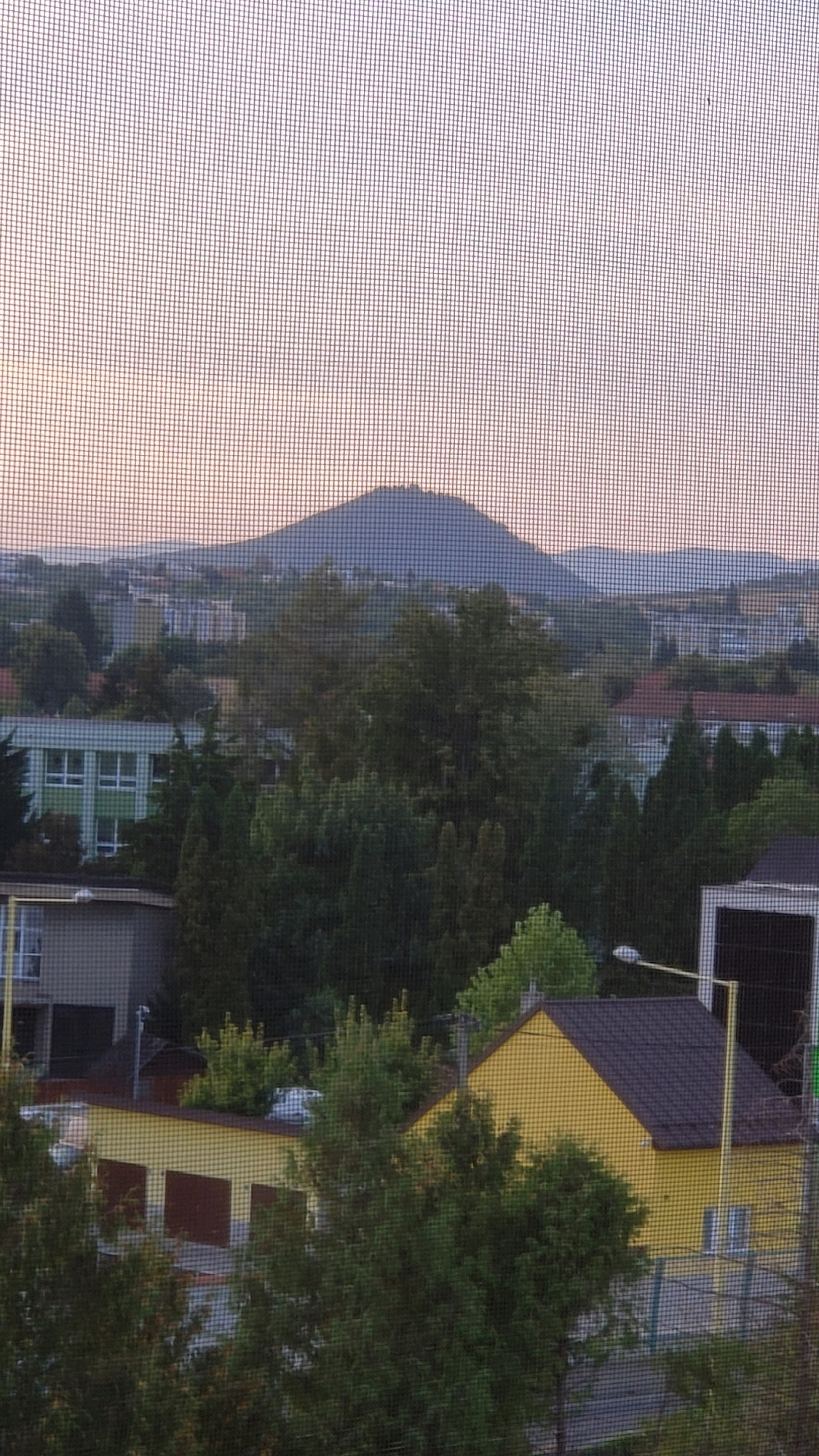 The image presents a view of a suburban or semi-urban landscape, dominated by a prominent conical hill in the background. The foreground is filled with trees and buildings. A light-colored building with green accents and a grid of windows is visible on the left. Adjacent to it is a gray structure. In the center and right of the foreground, a bright yellow building with a dark-tiled roof is prominent. This yellow building appears to be a garage or workshop, with three dark-brown garage doors visible. The scene is partially obscured by a window screen, creating a mesh pattern across the entire image. The lighting suggests it is either early morning or late evening, with a soft, diffused light coloring the sky and the distant hills. The location is identified as Prešov, Slovakia, and the prominent hill is likely the Zámčisko hill, a well-known landmark in the city, often referred to as "Spiš Castle" by locals due to its historical significance.