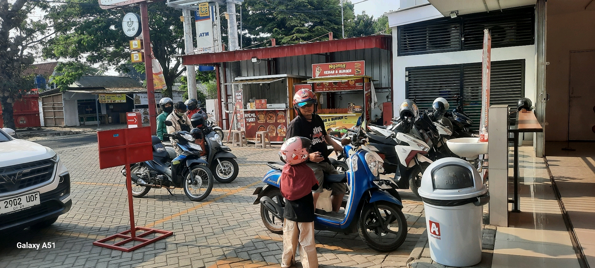 The image depicts an outdoor scene in front of a food stall in Wlingi, Indonesia. In the foreground, a white car with license plate B 2902 UOF is parked on a cobblestone street. To the right of the car, a red signpost with a red sign is visible. Several people on motorcycles are gathered, some wearing helmets. One person, a man wearing a black t-shirt and a red and white helmet, is seated on a blue scooter. Beside him, a child wearing a pink helmet is standing. Behind them, other motorcycles are parked. The food stall, identified as "Nyamill KEBAB & BURGER", has a red and white awning and displays pictures of food items. A white trash bin with the Alafamidi logo is situated to the right of the stall. In the background, trees and buildings can be seen, including a structure with the ATM logo and a sign that reads "Ja-di". The lighting suggests it is daytime.