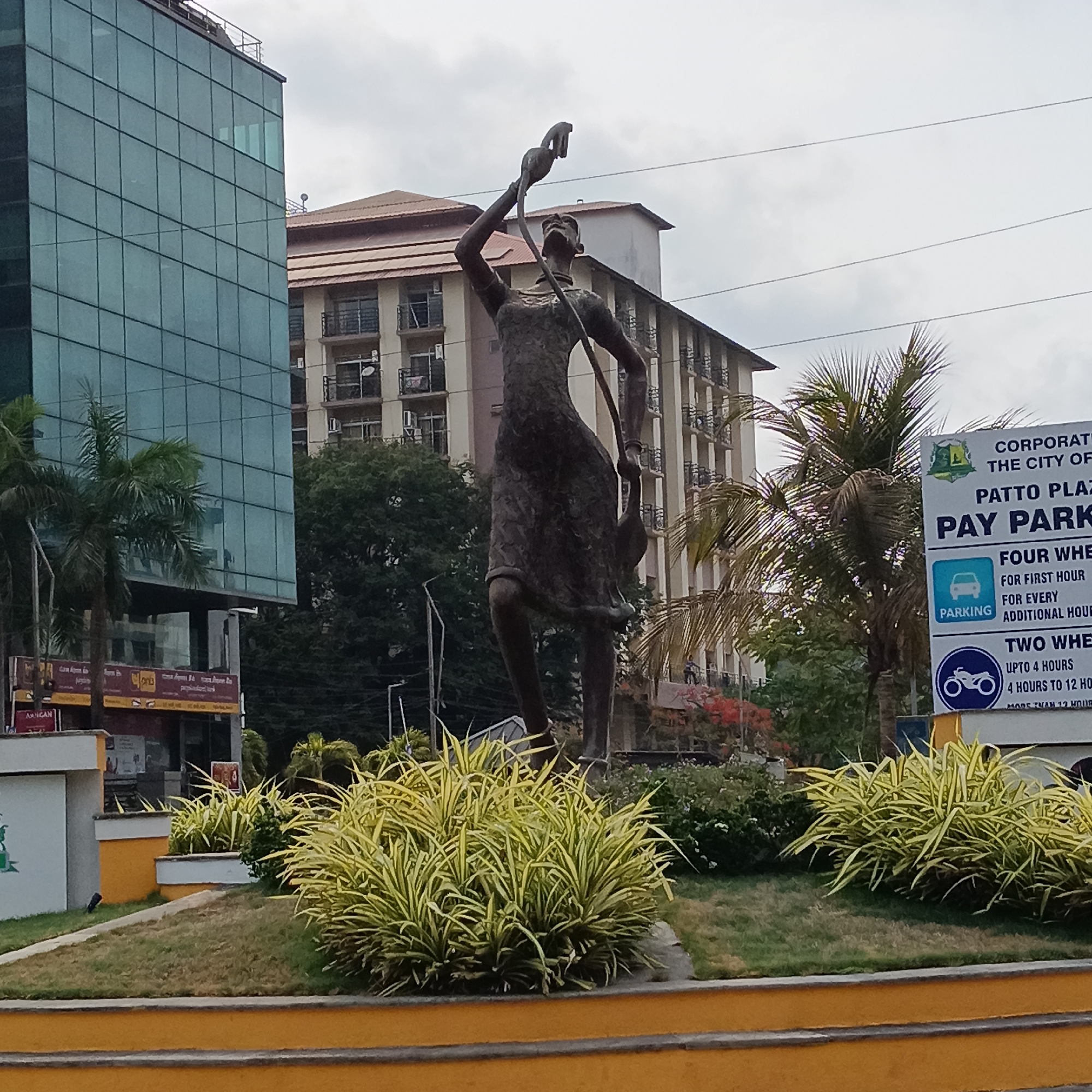 The image shows a bronze statue of a woman standing on a small, elevated, circular island surrounded by grass and plants in Panaji, India. The statue is situated in front of several buildings: a modern glass-fronted high-rise and a residential building with balconies. A sign for "Patto Plaza Pay Parking" is visible on the right. The sky is overcast.