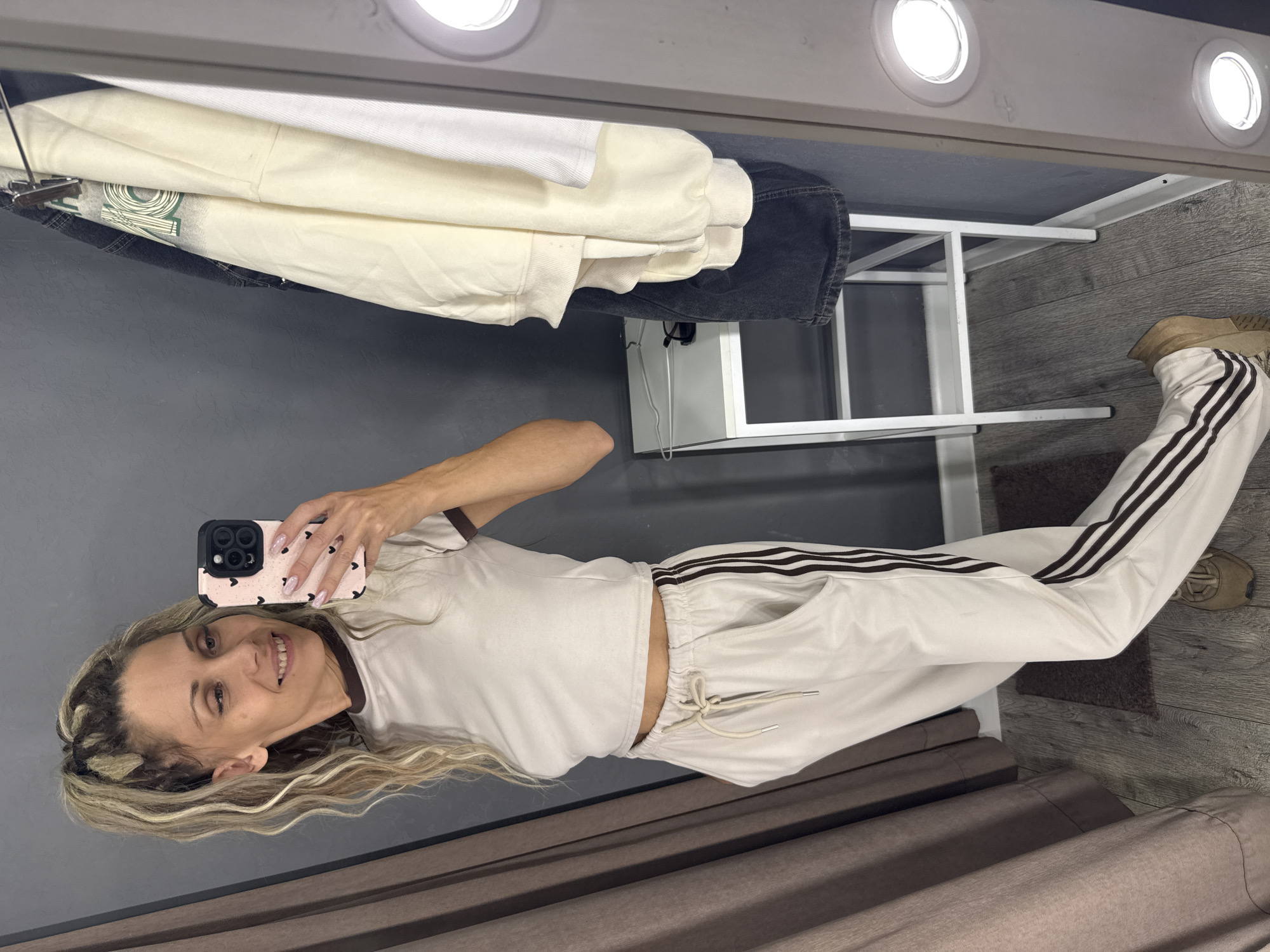 A woman is taking a selfie in a fitting room. She is wearing a light-colored crop top with a brown collar and matching light-colored sweatpants with brown stripes down the sides. She has blonde hair styled in a high ponytail and is holding a pink phone with a heart pattern. In the background, there are clothes hanging on a rack, a small white stool, and a large mirror with lights. The fitting room appears to be in a store.