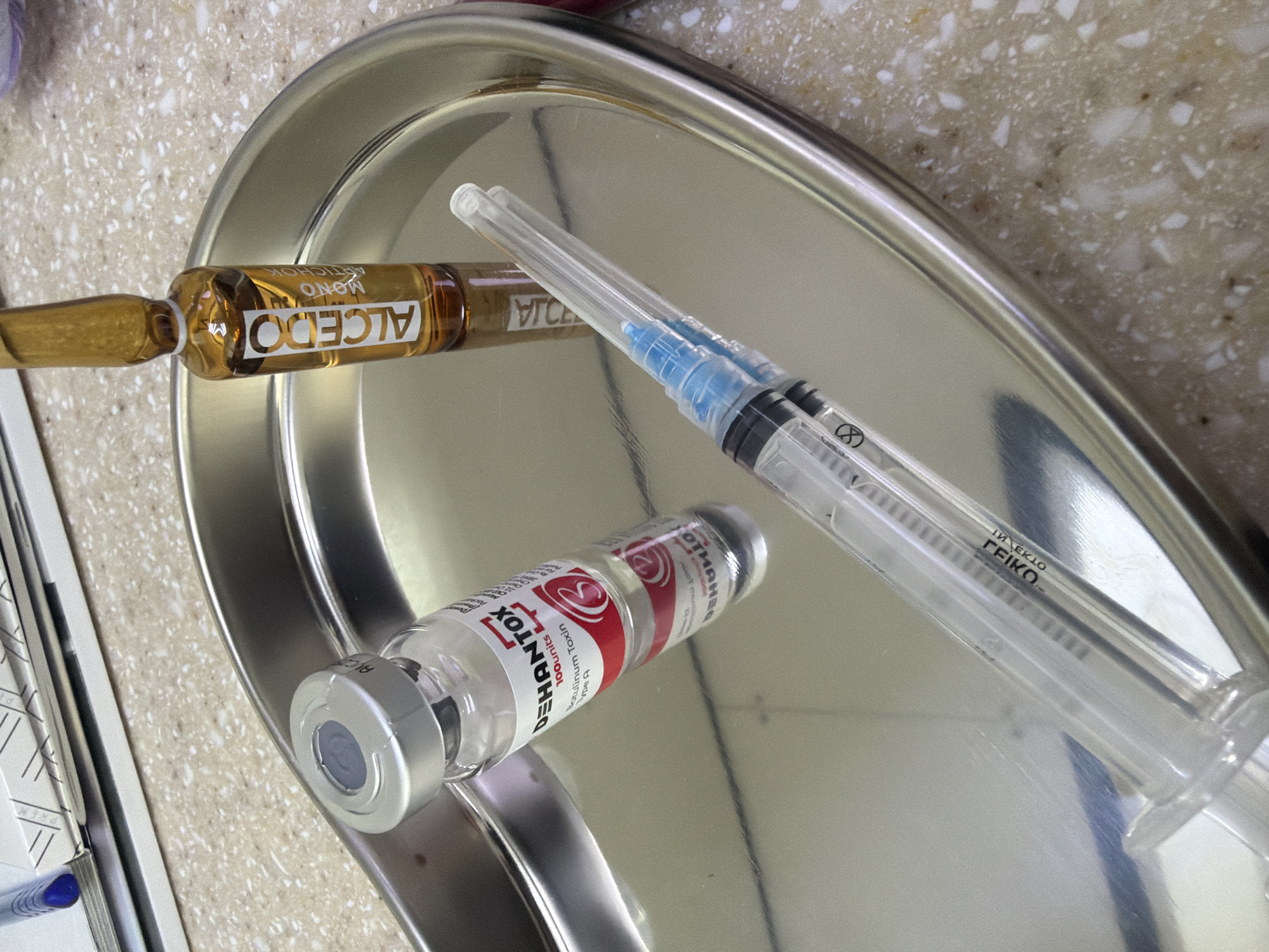 A top-down, slightly rotated, close-up shot shows a stainless steel tray with three items: a clear syringe, an amber ampule labeled "ALCEDO" with additional text "MONO ARTICHOK" printed vertically, and a clear vial labeled "DEHANTOX" with additional text "Botulinum Toxin Type A 100 units". The syringe is clear with a blue hub. The tray is positioned on a speckled surface with white, gray, and brown flecks. To the left, edges of white papers and blue ink are visible.