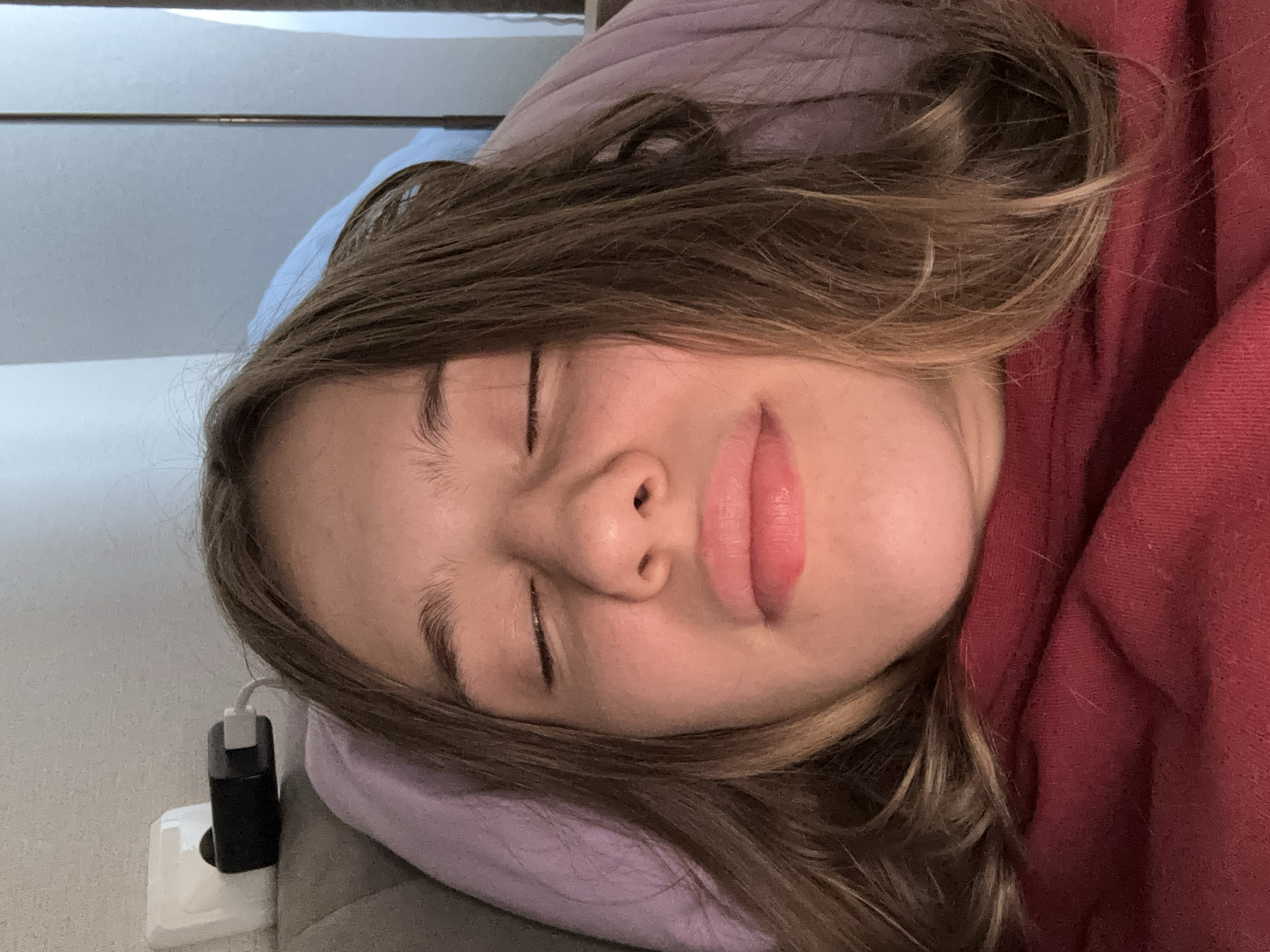 A young person with brown hair is lying down, with their eyes closed and head tilted back. Their face is illuminated by warm light, and their lips are slightly parted. They are wearing a maroon shirt, and a purple pillow is visible behind their head. In the background, a white electrical outlet with a black phone charger plugged in can be seen on a light-colored wall. The setting appears to be indoors, possibly a bedroom or living area.