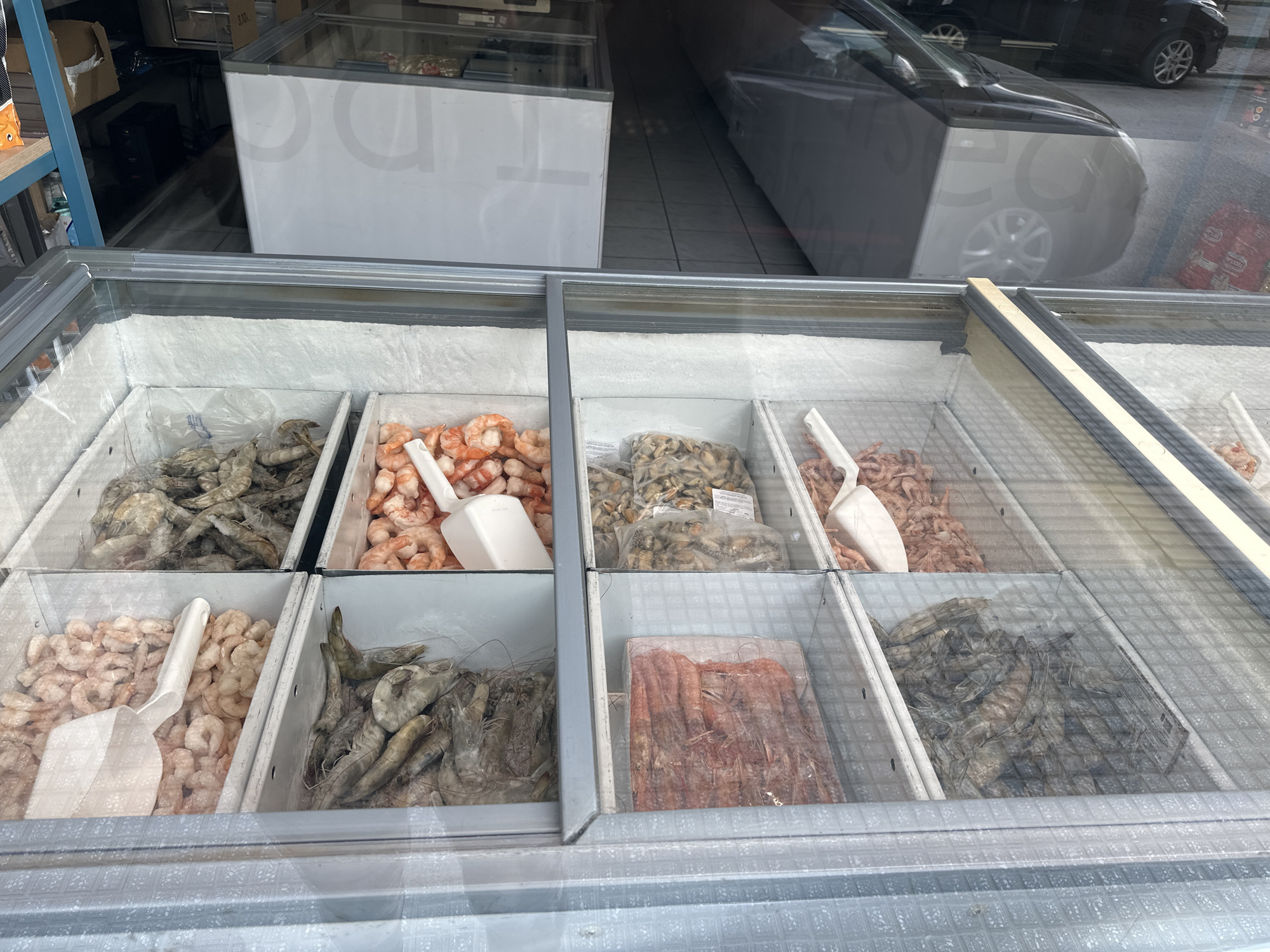 The image depicts a refrigerated display case filled with various types of shrimp in a store in Serres, Greece. The case is divided into several compartments, each containing a different variety of shrimp, including large grey shrimp, peeled pink shrimp, and smaller mussels in a clear packaging. A white plastic scoop is present in some of the compartments. The freezer case is visible through a window, and the reflection shows a car parked on the street.