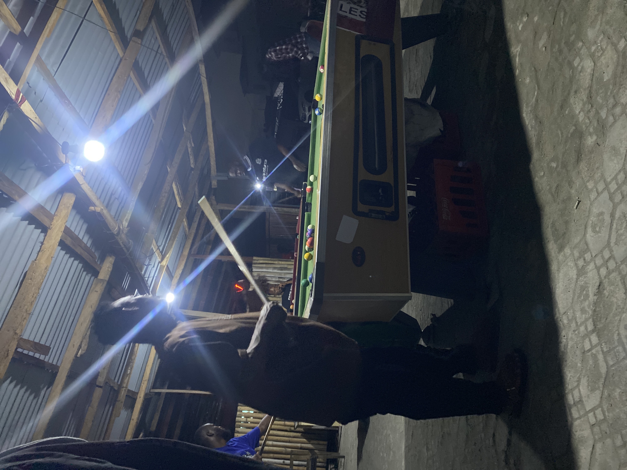 The image depicts a dimly lit indoor or covered outdoor space at night in Ado, Nigeria, characterized by a utilitarian construction with a corrugated metal roof supported by visible wooden beams. Bright artificial light sources hang overhead, generating significant lens flares and illuminating parts of the scene while casting deep shadows elsewhere.
The central activity revolves around a standard green-felted pool table, where several individuals are gathered. In the foreground, a man in dark clothing, possibly holding a pool cue, stands observing the table, his head silhouetted against a bright light source. Another person in a blue shirt is partially visible towards the bottom right, also seemingly engaged in the game. Further in the background, near the other end of the pool table, more figures are present, including one wearing a plaid shirt, suggesting a social gathering centered on playing billiards.
The pool table has various colored balls scattered on its surface, and the side of the table features the partial text "LES". On the patterned concrete floor, a red crate with "Coca-Cola" branding is visible. The bright lights cast prominent, elongated shadows across the floor and surrounding areas. The scene conveys a casual, late-night atmosphere in an informal setting.