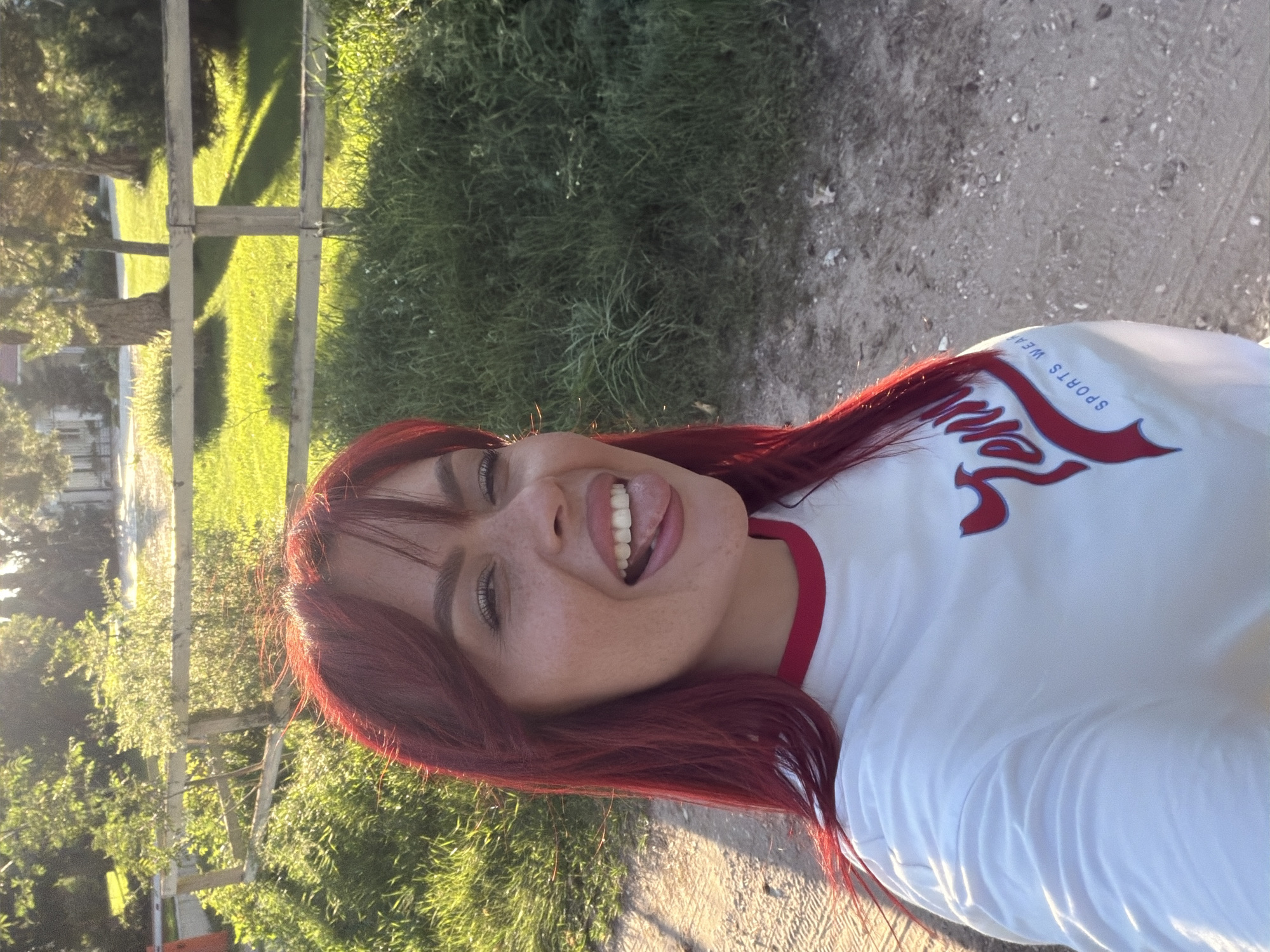 The image is a selfie taken outdoors. A person with long, bright red hair and freckles is in the foreground, sticking their tongue out and smiling. They are wearing a white t-shirt with red trim and a red logo on it that appears to say "TENNI SPORTS WEAR". The background shows a wooden fence, green grass, and trees, illuminated by the setting sun, suggesting a rural or park-like setting. The ground in the foreground is dirt or gravel. The overall impression is of a casual, candid photograph taken during golden hour. The location is identified as Узюково, Russia.