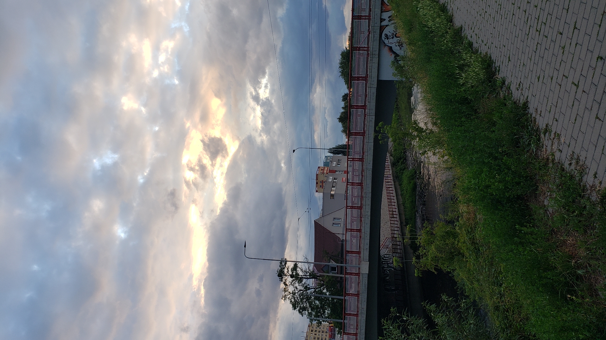 The image captures a view of the river Hornád in Prešov, Slovakia, near the Slánska street bridge. The sky above is filled with dramatic, heavy clouds, with patches of bright sunlight breaking through, suggesting either dawn or dusk. A multi-lane bridge with red railings spans the river. Below the bridge, the river flows, with the riverbanks lined with lush green vegetation. A paved pedestrian walkway is visible on the right side of the image, with the same lush vegetation bordering it on the left. Buildings, including what appears to be a residential building with a red roof and other larger structures with signage, are visible beyond the bridge and tree line. Streetlights are present on the bridge. Graffiti is visible on the underside of the bridge.