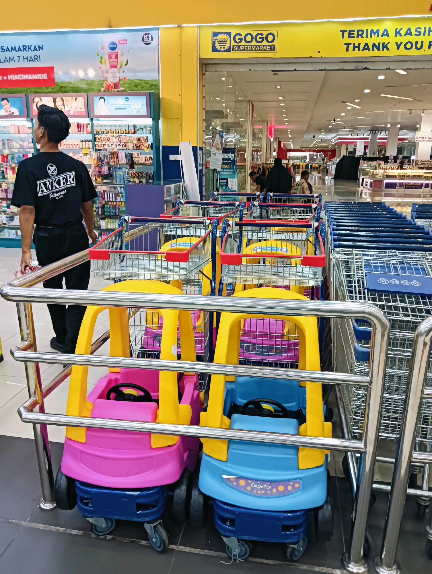 The image depicts the entrance area of a GOGO Supermarket, likely in Batam, Indonesia, identifiable by the signage. In the foreground, a metal barrier separates the viewer from two brightly colored children's shopping carts, one pink and yellow, the other blue and yellow. These carts are designed to resemble small cars with steering wheels. Behind the barrier and to the left, a young man wearing a black t-shirt with an "ANKER" logo and black pants stands facing away from the camera, holding a white item in his left hand. He appears to be an employee, possibly guiding shoppers or managing the carts. In the background, rows of standard shopping carts are visible, some made of blue plastic and others of silver metal. The supermarket interior is visible beyond the carts, with shelves stocked with various products, including toiletries and cosmetics, and overhead signage indicating promotions. The overall environment suggests a retail setting with customers present but not prominently featured.