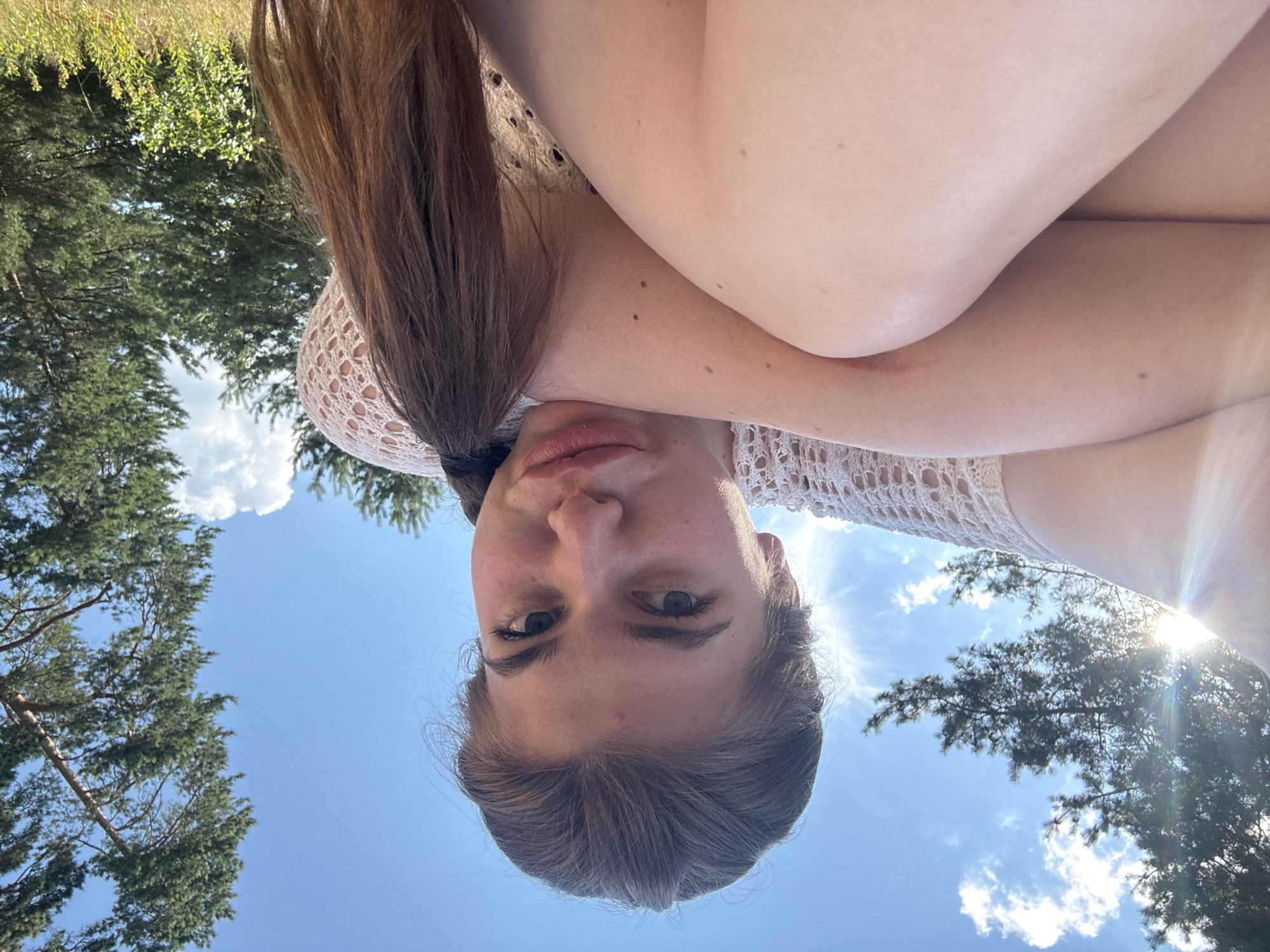 In a low-angle, outdoor shot, a young woman with light brown hair tied in a ponytail poses in a natural setting. She is wearing a cream-colored crocheted top. Her arms are crossed, and she is looking directly at the camera. The background consists of a clear blue sky with scattered white clouds and tall pine trees. The sun is shining brightly, creating a lens flare effect on the left side of the image. The scene appears to be in a wooded area, possibly a park or forest, consistent with the environment of Kurovskoye, Russia.