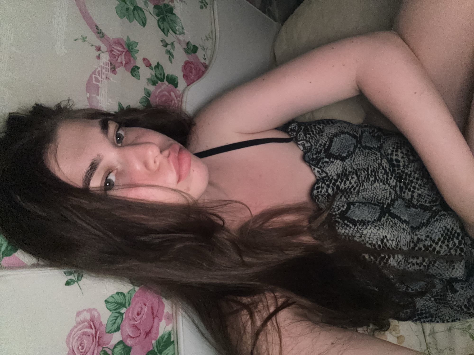 A young woman with long, dark hair is lying on her side, facing the camera. Her head is tilted back slightly, and her eyes are looking directly at the viewer. She is wearing a black bra with a snake-print top over it. The background appears to be a floral-patterned fabric, possibly upholstery or bedding, with pink roses and green leaves. The lighting suggests it might be nighttime or a dimly lit room. The woman's expression is relaxed and casual. There are no visible signs of text in the image. The location context is Мещёры, Russia.