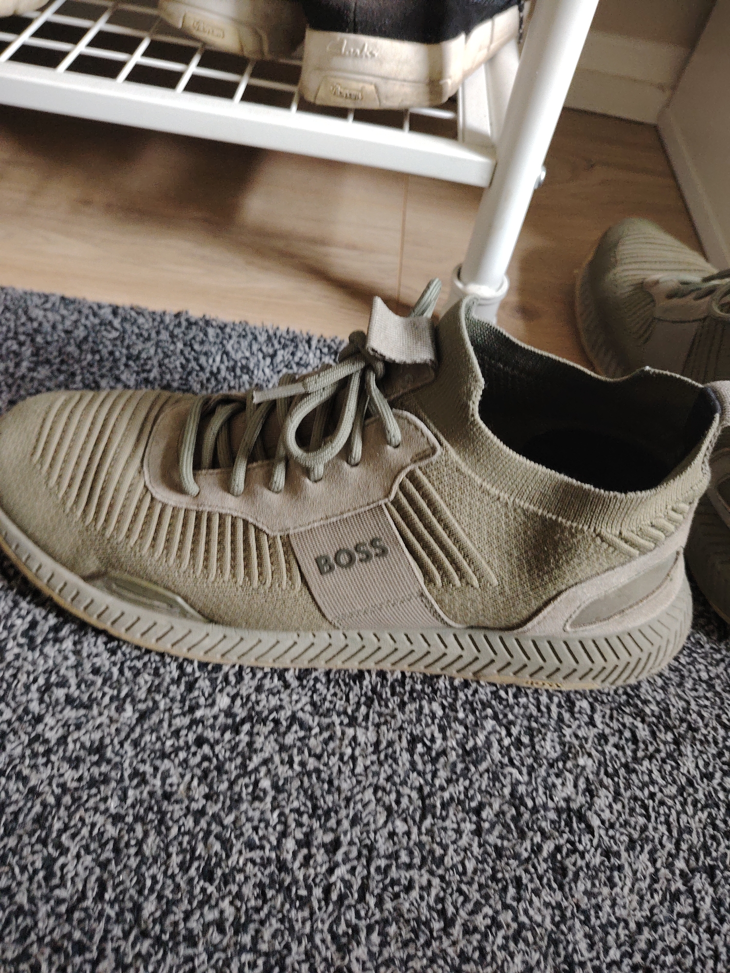The image displays an olive-green BOSS brand sneaker, specifically the left shoe, positioned on a mottled grey and black carpet. The sneaker features a knitted upper with prominent vertical ribbed textures on the toe box and lateral panels. A flat fabric overlay accommodates a lacing system with matching olive-green laces. The word "BOSS" is debossed in dark capital letters on a rectangular fabric band located on the lateral side of the shoe. The sole is thick, uniformly olive-green, and exhibits a pronounced chevron/zigzag tread pattern.
In the mid-ground, a light brown wooden floor is visible behind the carpet. A white metal shoe rack stands on this wooden floor, with one vertical support pole clearly visible. On the uppermost shelf of this rack, two pairs of light-colored shoes, one with a visible "Clarks" brand logo, are present. A second olive-green sneaker, partially obscured and likely the right shoe, is situated on the wooden floor in the upper right quadrant of the image. The scene is an indoor environment located in Huntingdonshire, United Kingdom.