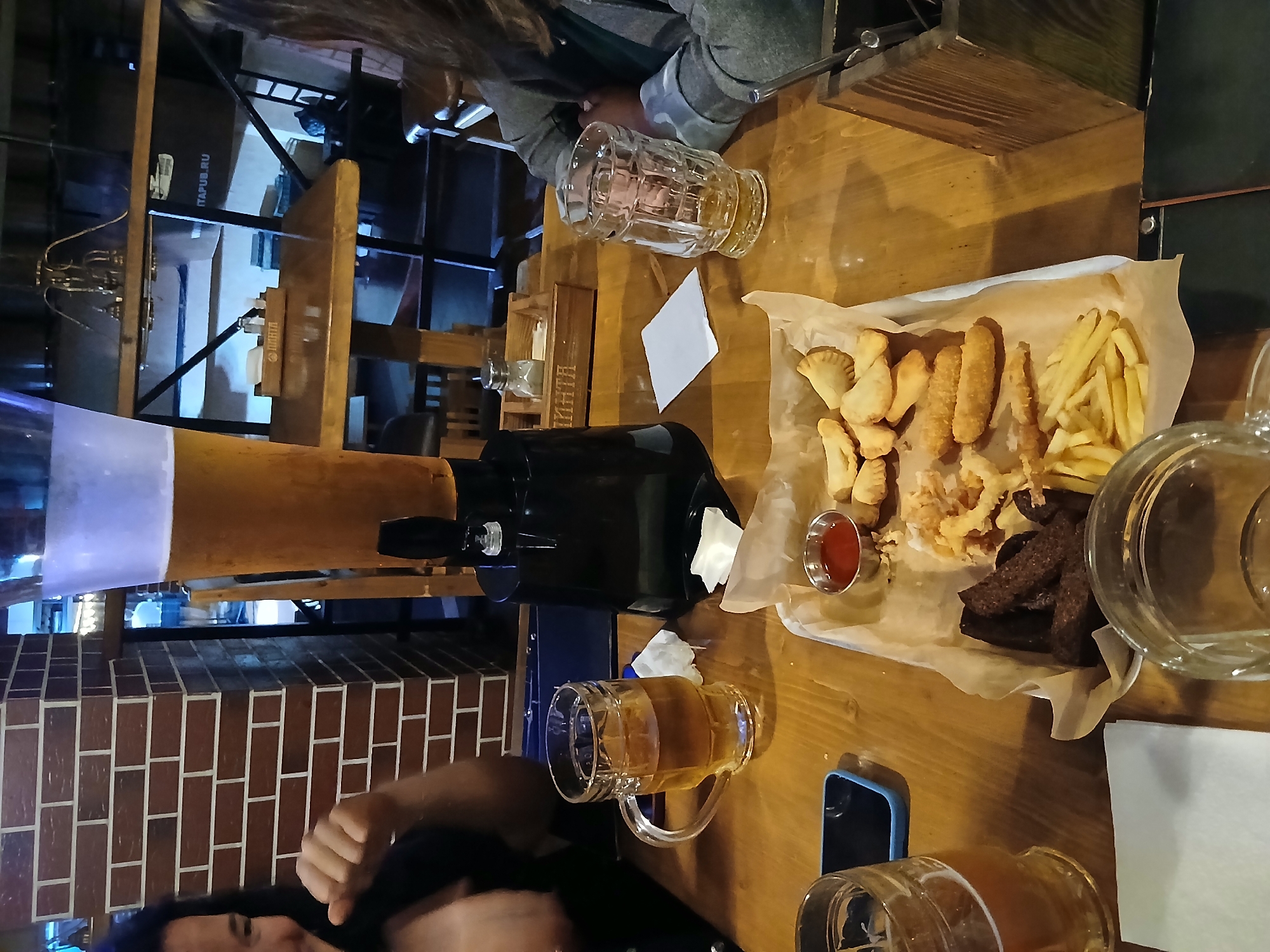 The image depicts a group of people enjoying drinks and food at a table in what appears to be a pub or restaurant. A large beer tower is prominently displayed on the table, along with several glasses of beer, a plate of fried appetizers including french fries and breaded items, and a small dish of ketchup. A person is visible in the foreground on the left, and another person's arm is visible on the right. The setting features wooden tables and brick walls, suggesting an indoor establishment. While the image does not explicitly state the location, it is stated to be in the city of Izhevsk, Russia. The overall scene suggests a casual social gathering.
