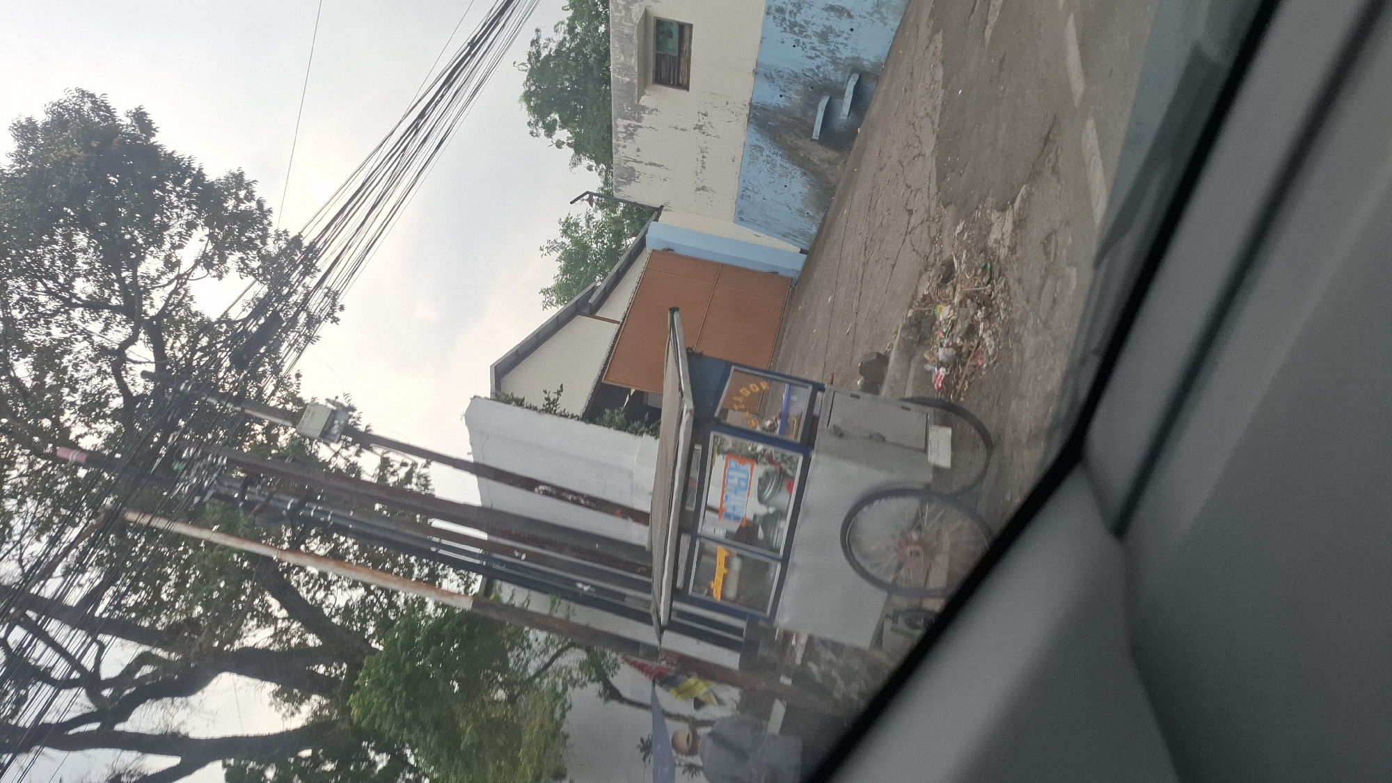The image depicts an outdoor scene in Cilaku, Indonesia. A food cart with a blue and silver exterior and a metal canopy is parked on a cracked asphalt surface. The cart has two wheels, one of which is partially visible and appears to have red spokes. A person in a grey jacket and a cap is walking away from the cart towards the left side of the frame. Behind the cart, a tall, light-colored structure with a sloped roof is visible. To the right of this structure is a large orange gate. Further to the right, a white wall with blue accents runs along the side of a building. Overhead, a dense network of electrical wires crisscrosses the frame, supported by utility poles. Trees with lush green foliage are also present, contributing to the urban yet somewhat overgrown environment. A pile of trash is situated near the food cart on the ground. The sky is overcast, suggesting cloudy weather.