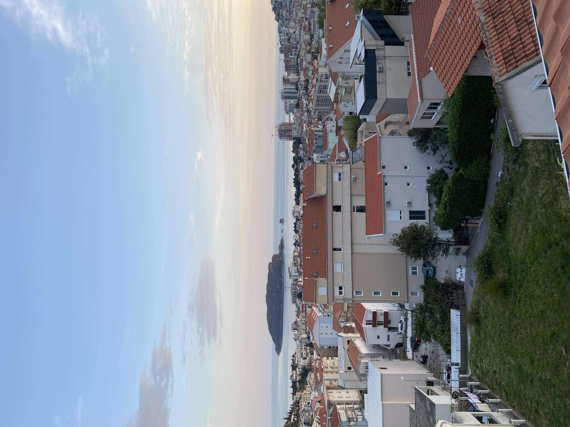 Here is a description of the image:
The image depicts a coastal cityscape, most likely Budva, Montenegro, during either sunrise or sunset. The sky is a gradient of light blue and pale orange, indicating the time of day. The sea is calm and stretches to the horizon. A peninsula or island is visible in the distance, jutting out into the sea.
The foreground consists of residential buildings with terracotta tile roofs, a common architectural feature in this region. The buildings appear to be a mix of older and more modern structures, densely packed together. Green vegetation is interspersed among the buildings, including trees and grassy areas. A small road or path is visible between some of the buildings. There are cars on the street and clothes are hanging outside.