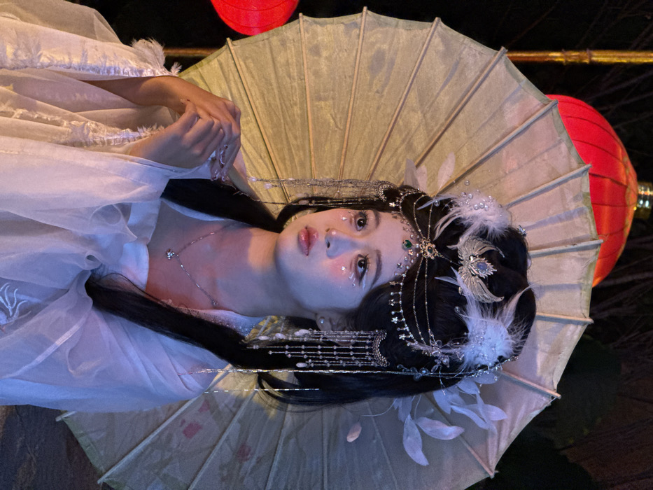 A young East Asian woman is prominently featured in a portrait-style image, captured at night in Yuzhong District, China. She is positioned slightly to her right, with her head turned to the left and her gaze directed upwards and to the left. Her dark hair is meticulously styled in an elaborate updo, adorned with a complex silver-colored headpiece that includes white feathers, a central bird-like motif with a blue gemstone, and draped chains featuring small stars and beads across her forehead and temples. Long, multi-strand tassels with white, pearl-like beads cascade from the sides of her head to her shoulders. Her face is made up with white highlights on her lower eyelids and small star-shaped embellishments on her cheekbones, complementing her glossy, slightly parted lips.
She wears a traditional Hanfu-style garment, light-colored and seemingly made of sheer, flowing fabric, with faint embroidery visible on the sleeves. A delicate silver-colored chain necklace with a small pendant is visible around her neck. Her hands are gently clasped in front of her chest, exhibiting ornate, long finger extensions or decorative nail art. Directly behind her head, a large, pale yellow or light green traditional Chinese oil-paper umbrella is held open, displaying its wooden ribs. In the blurred background, two illuminated red Chinese lanterns are visible: one large at the upper left and a smaller one positioned to the lower left, casting warm red light. The overall scene is dimly lit by the lanterns and ambient sources, creating a contrasting interplay of warm red and cooler blue-purple tones on the subject.