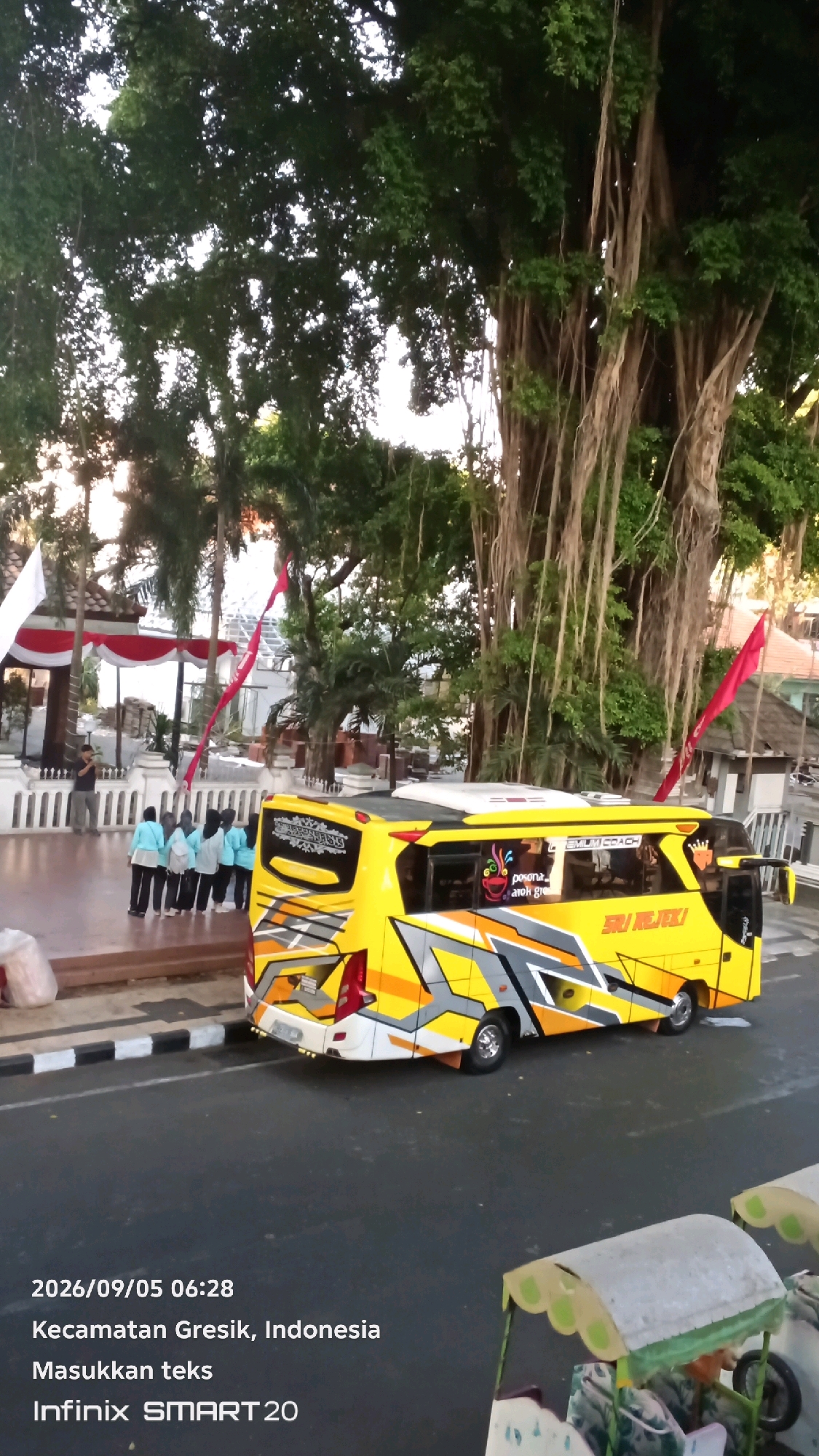 In Gresik, Indonesia, a vibrant yellow bus with a distinctive graphic design is parked on a street. The bus is adorned with intricate patterns in gray and orange, and features text that reads "SRI REJEKI" on its side. In the background, a group of people, primarily women wearing light blue tops and dark pants, stand near a white building with a red and white striped awning. They appear to be in a leisurely setting, possibly near a park or public space, as indicated by the presence of large trees with hanging aerial roots and a decorative white fence. The scene is captured with a timestamp of "2026/09/05 06:28" and the device information "Infinix SMART 20".