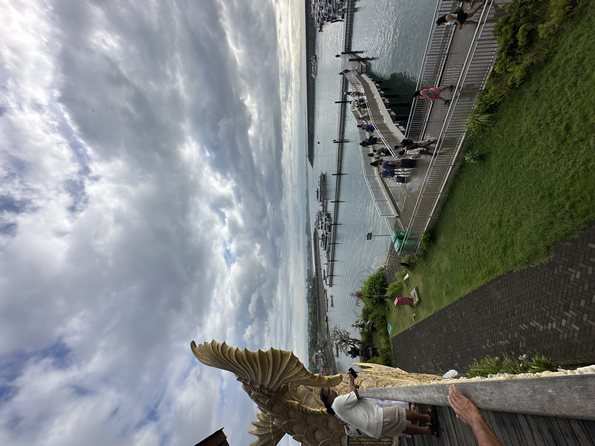 The image shows a view of a harbor from a high vantage point, likely in Denpasar, Indonesia. In the foreground is a large, golden statue, possibly of a fish or mythical creature, which appears to be part of the railing or structure where the picture is taken. A person in a white shirt and tan shorts is standing next to the statue, holding a camera and seemingly taking a picture of the scene. In the background, there is a pier with people walking on it, along with several boats and yachts docked in the harbor. Beyond the harbor, the coastline and some buildings can be seen. The sky is overcast with cloudy skies. Part of a brick walkway and green grass are visible in the mid-ground, along with a metal railing.