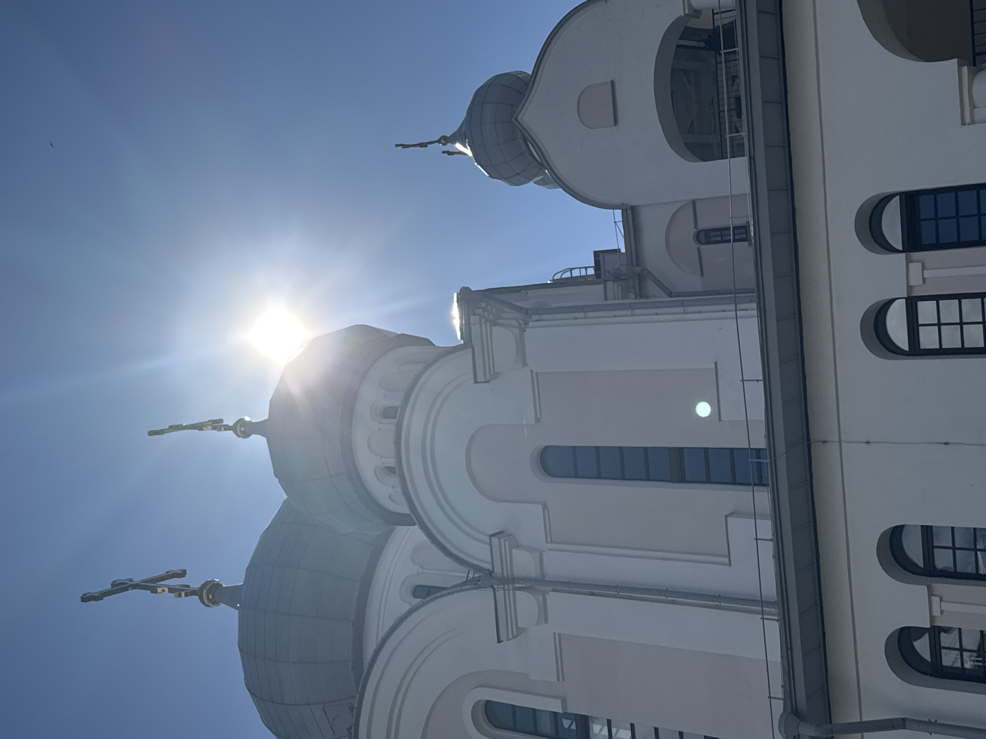 The image shows a low-angle view of the domes and facade of a church, likely an Orthodox Christian church, under a bright, sunny blue sky. The sun is prominently visible, creating a lens flare effect. Several domes, topped with crosses, are visible, with the largest dome in the foreground. The church is constructed with light-colored materials, likely stucco or stone, with arched windows and decorative architectural elements. The style suggests a classic Russian Orthodox design. While no specific location in Saint Petersburg is identified, the architecture is consistent with many churches found in the city. There are no people or explicit actions depicted. The environment is clearly outdoors, with the clear sky and sunlight indicating fair weather.