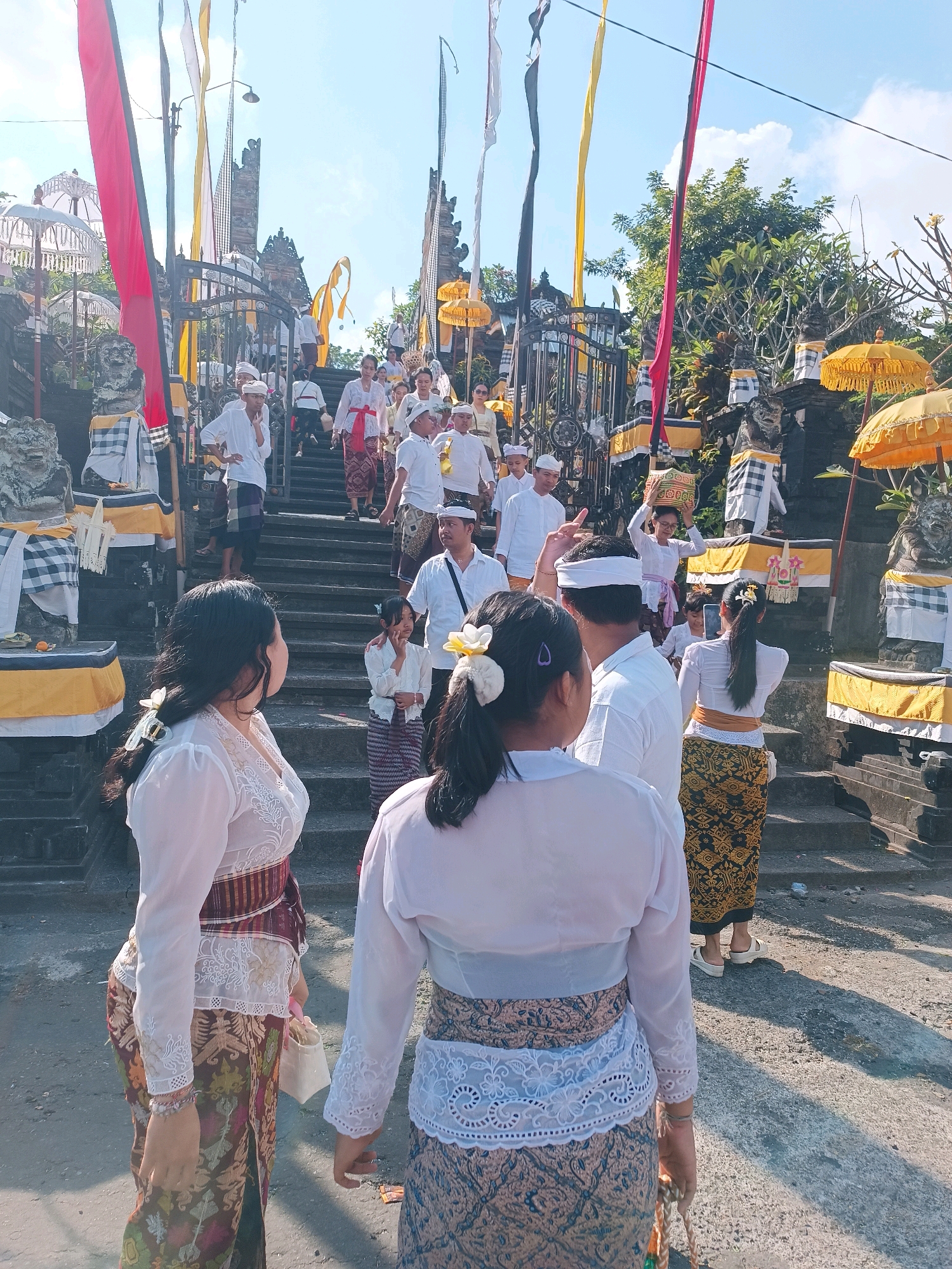In Tabanan, Indonesia, a group of people are ascending a stone staircase towards a temple complex. Many are dressed in traditional Balinese attire, featuring white tops and patterned sarongs. Some individuals are holding offerings or religious items. The scene is adorned with colorful flags and yellow umbrellas, typical of ceremonial decorations. Statues and ornate stone carvings flank the staircase, suggesting a significant cultural or religious event. The lighting indicates it is daytime, under a bright sky.