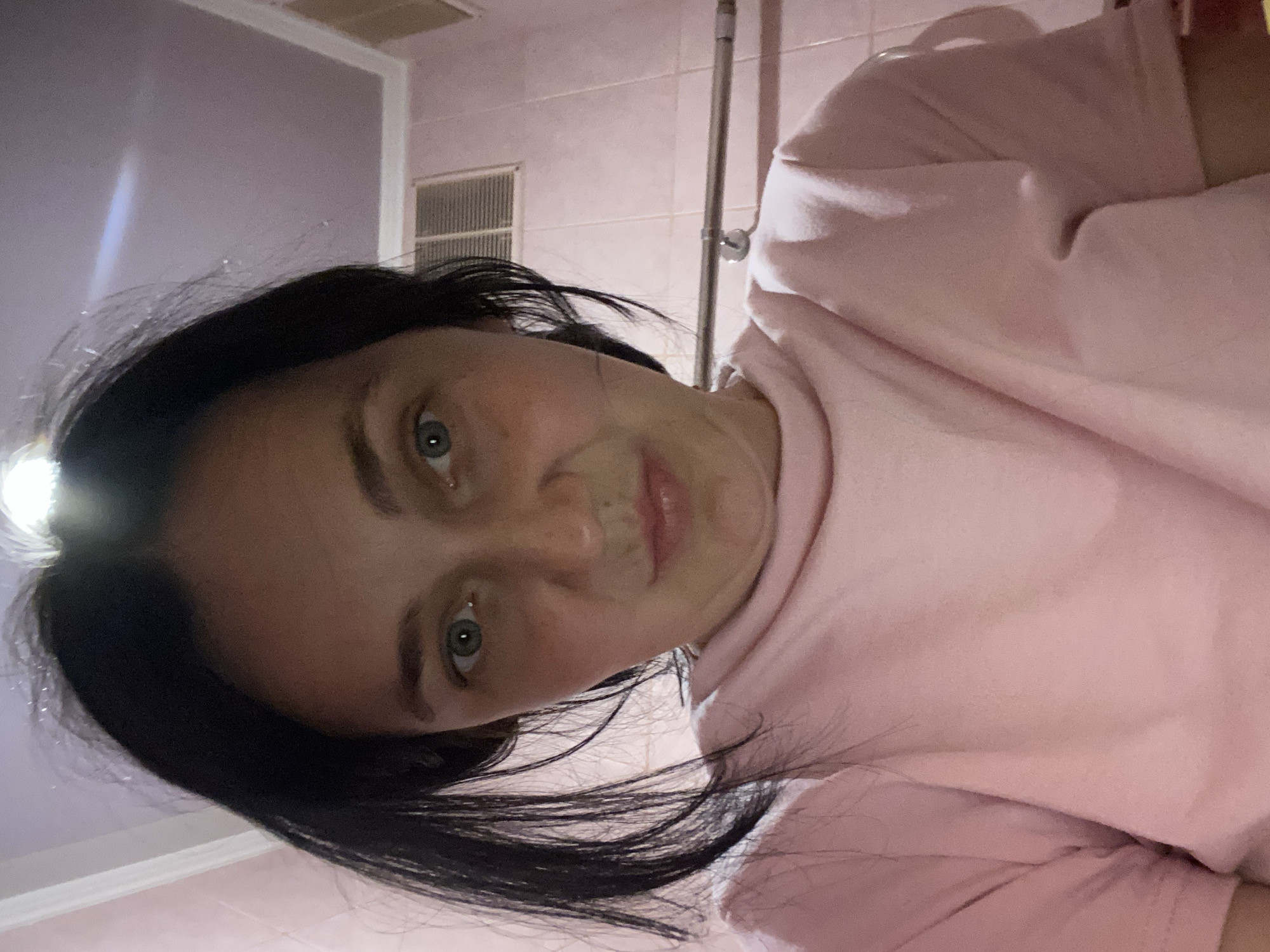 A young woman with dark hair and blue-green eyes is taking a selfie. She is wearing a pink t-shirt and is positioned in front of a pink tiled wall. The room appears to be a bathroom, with a vent and a towel rack visible in the background. The lighting is overhead and seems to be causing a glare on the ceiling. The woman's expression is neutral, with her lips slightly pursed.