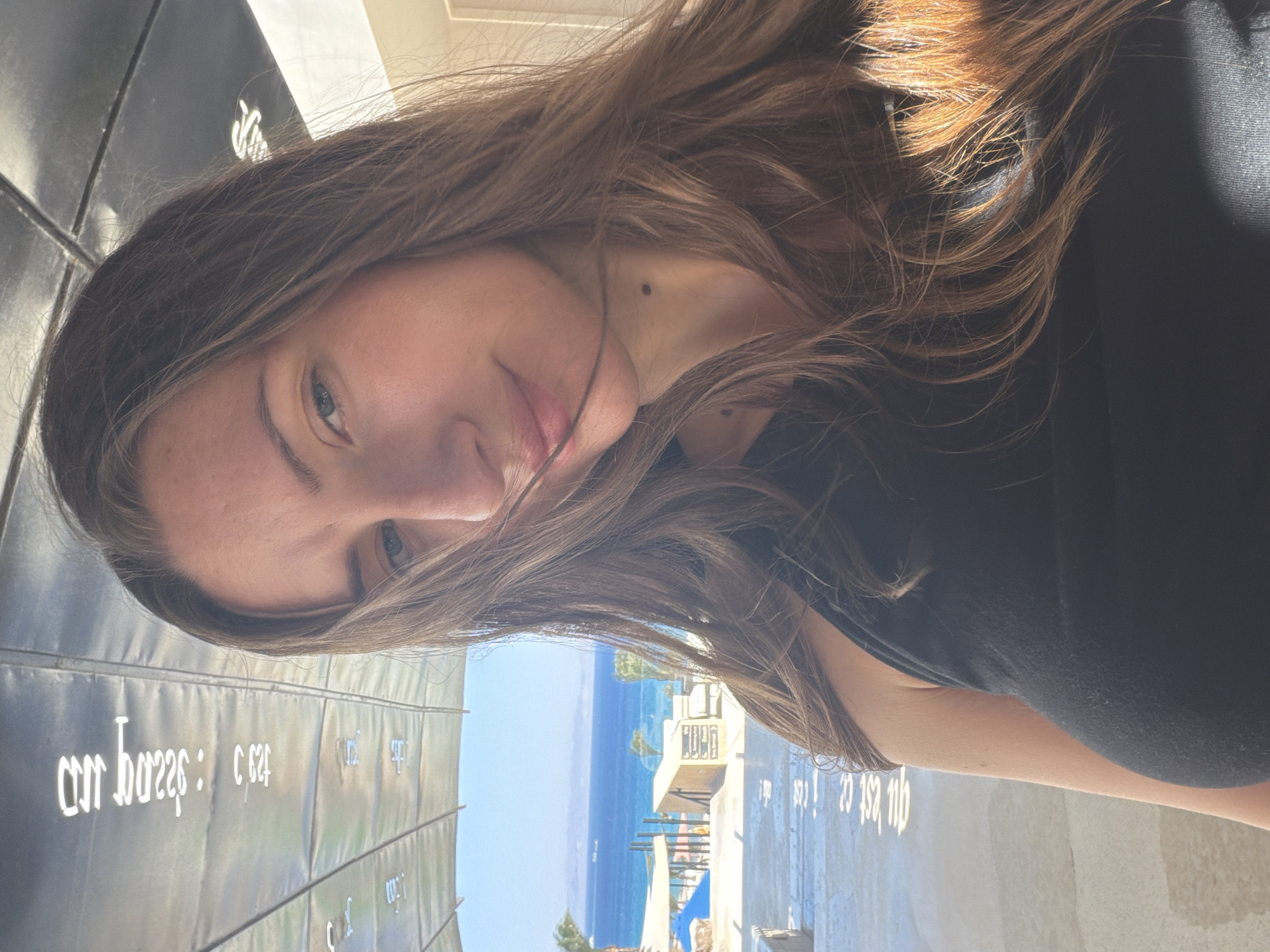 A close-up, front-facing selfie of a young Caucasian woman with long, light brown hair is captured outdoors. She is wearing a black tank top and has a faint smile. Her gaze is directed slightly upwards and to the left. The background reveals a body of water, likely the Red Sea, under a bright blue sky, with a sandy area, some palm trees, and structures in the distance, suggesting a resort or beach location in Dahab, Egypt. A metallic structure with reflective panels is visible overhead, and text in an Asian script is printed on the floor in the foreground.