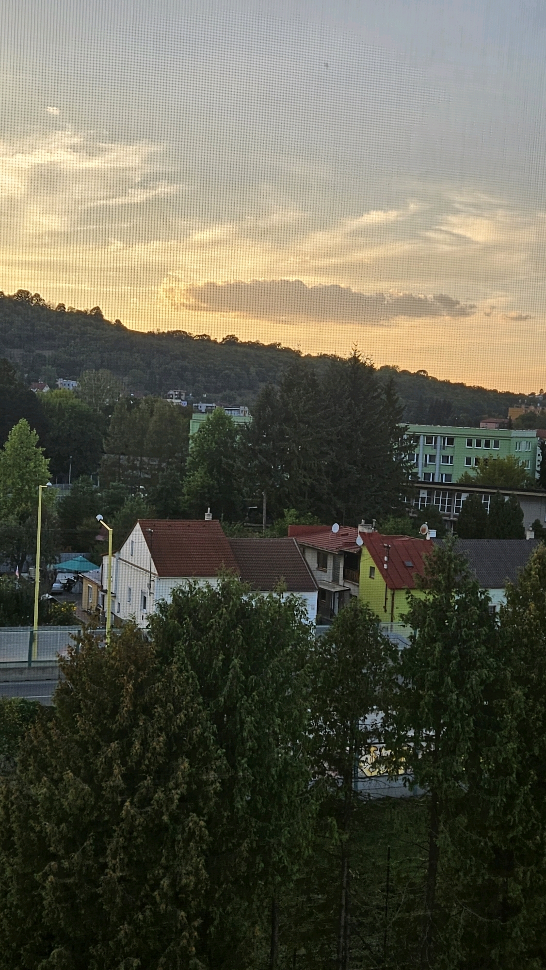 The image shows a view of a residential area with houses, trees, and a large green building in the background, likely taken from an elevated position looking towards a hillside at dusk. The sky is a pale orange and yellow, indicating the setting sun, with some scattered clouds. In the foreground, there are several trees. Further back, there are houses with red and brown roofs, and a bright yellow house. A road and a fence are visible to the left of the houses. The large green building in the distance appears to be an apartment complex or an institutional building. The scene is likely captured from a window, as a grid pattern, possibly from a screen or a window pane, is visible across the entire image. This view is consistent with a residential area on the outskirts of Prešov, Slovakia, possibly near the Sekčov housing estate, given the presence of large apartment blocks and surrounding greenery.