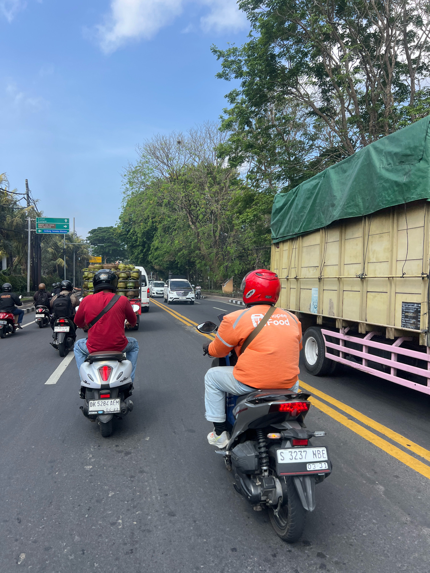 The image shows a street scene in Denpasar, Indonesia, on a clear day. A multi-lane road is filled with traffic, predominantly motorcycles. In the foreground, two individuals are riding motorcycles. The person on the left is wearing a red shirt and blue jeans, while the person on the right is wearing an orange jacket with "Shopee Food" branding and light blue jeans. Both are wearing helmets. To the right of the motorcycles, a large yellow truck with a green tarp covering its cargo is traveling in the same direction. In the background, several cars and other motorcycles are visible, along with trees and a blue sky with scattered clouds. Traffic signs are present on the left side of the road, indicating directions to various locations. The road is marked with yellow and white lines.