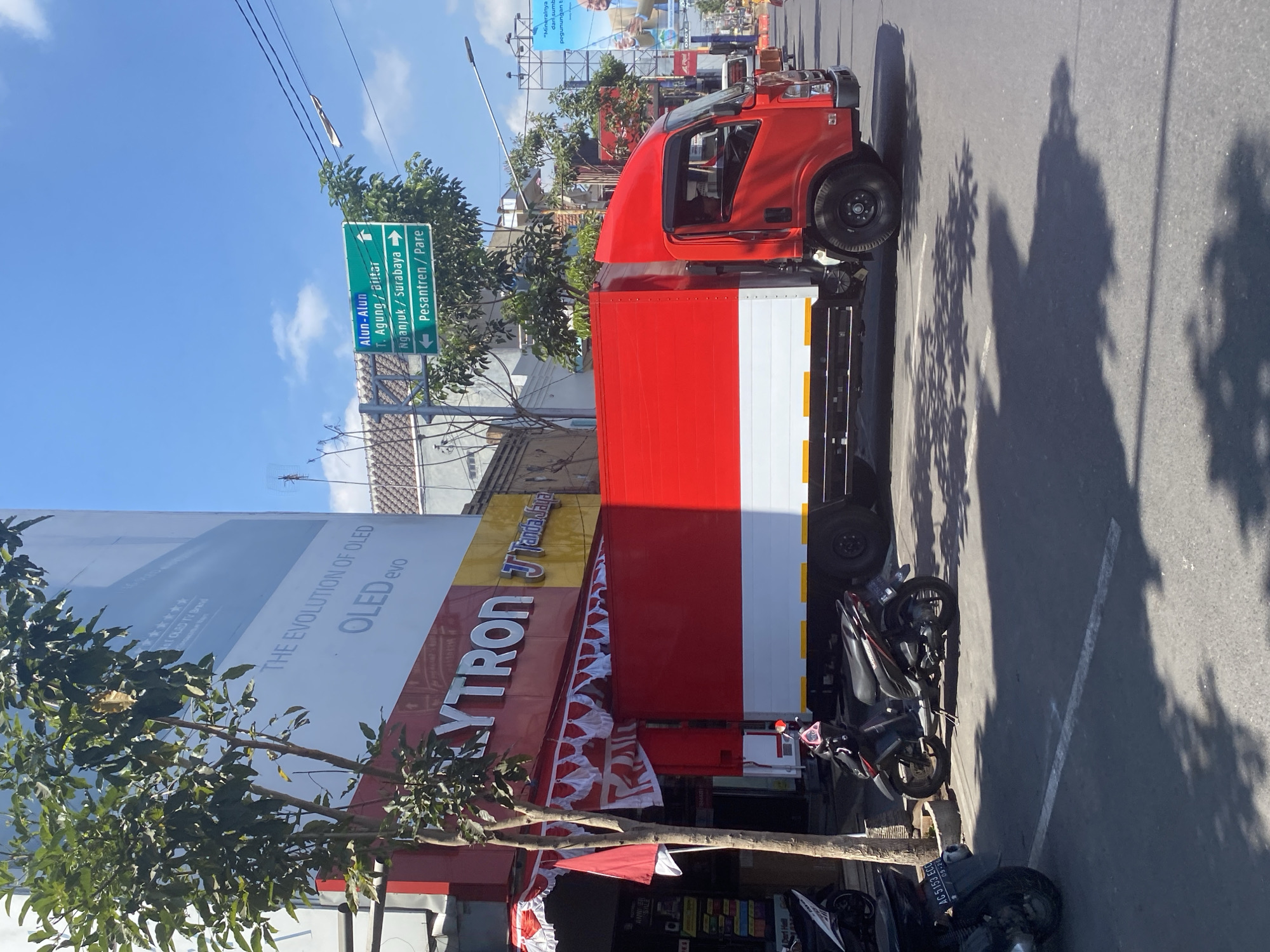 In Kediri City, Indonesia, on a bright, sunny day, a red and white delivery truck is parked on the side of a street. The truck's cargo area is painted in horizontal stripes of red and white, resembling the Indonesian flag. To the left of the truck is a store with a red awning and prominent "POLYTRON" signage. In front of the store, a tree stands tall, and two motorcycles are parked. A street sign in the background indicates directions to Alun-Alun, T. Agung/Blitar, Nganjuk/Surabaya, and Pesantren/Pare. The sky is a clear blue with a few scattered clouds. The scene captures a typical urban streetscape in Indonesia, with a focus on the vibrant colors of the truck and the store.