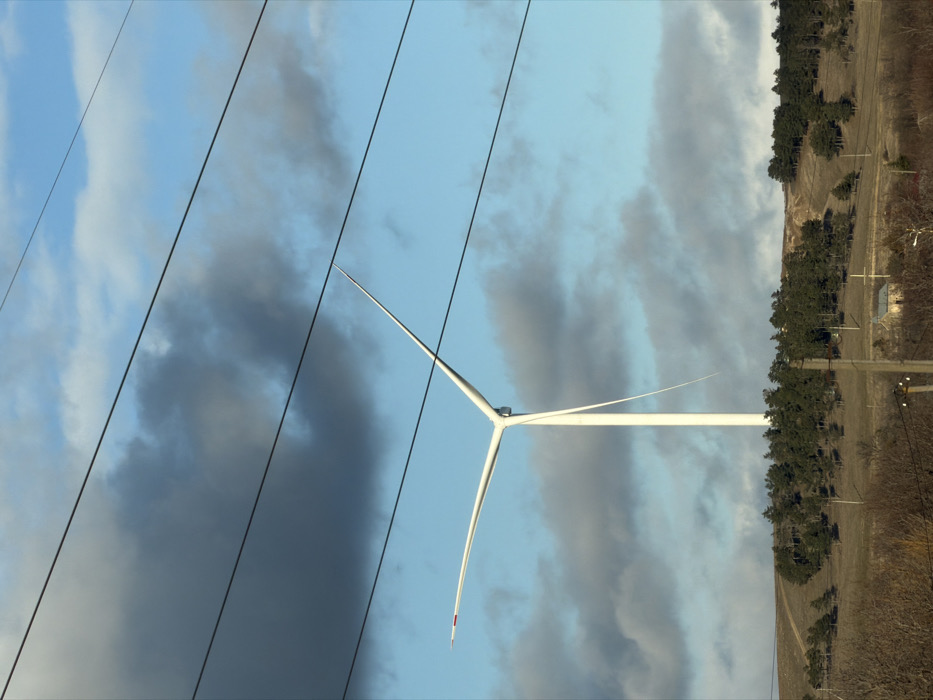 The image depicts an outdoor scene in the city of Ruisi, Georgia, under a partly cloudy sky. A large, white, three-bladed wind turbine is the central subject, positioned in the middle ground with its blades stationary. One blade extends horizontally to the left, another points upwards and to the right, and a third points downwards and to the right; all blade tips feature small red markings. Multiple dark power lines, approximately four to five, traverse horizontally across the upper portion of the frame, partially obscuring the sky and the turbine blades. The sky transitions from light blue to a darker grey with interspersed white and grey clouds. The lower third of the image features a rolling, hilly landscape with sparse, dry brown vegetation and clusters of dark green evergreen trees. A dirt path is visible ascending a distant hill to the left of the turbine. In the lower-right mid-ground, a small, light-colored building with a dark roof is partially visible behind a utility pole. No people or active human interactions are present.