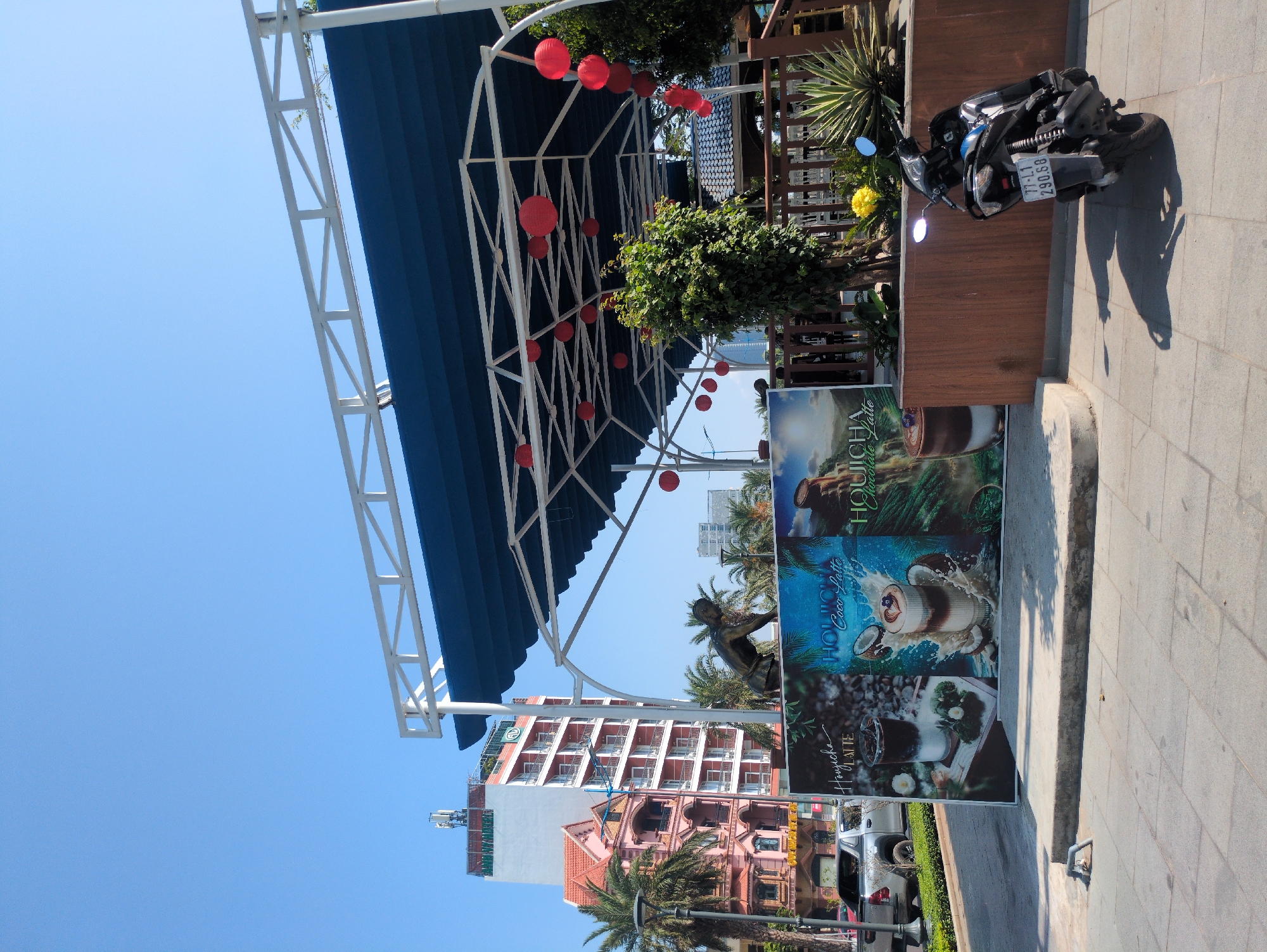 The image captures a street scene in South Quy Nhon Ward, Vietnam. A blue awning supported by white metal framework is strung with red lanterns. Adjacent to this is a wooden structure with plants and a parked black scooter bearing the license plate 77-L1 290.68. In the foreground are three advertising posters, displaying images of beverages and the name "Houicha". In the background, buildings are visible, including a red-and-white high-rise. The sky is clear and blue. The scene is captured from a slightly low angle. The pavement appears to be tiled.