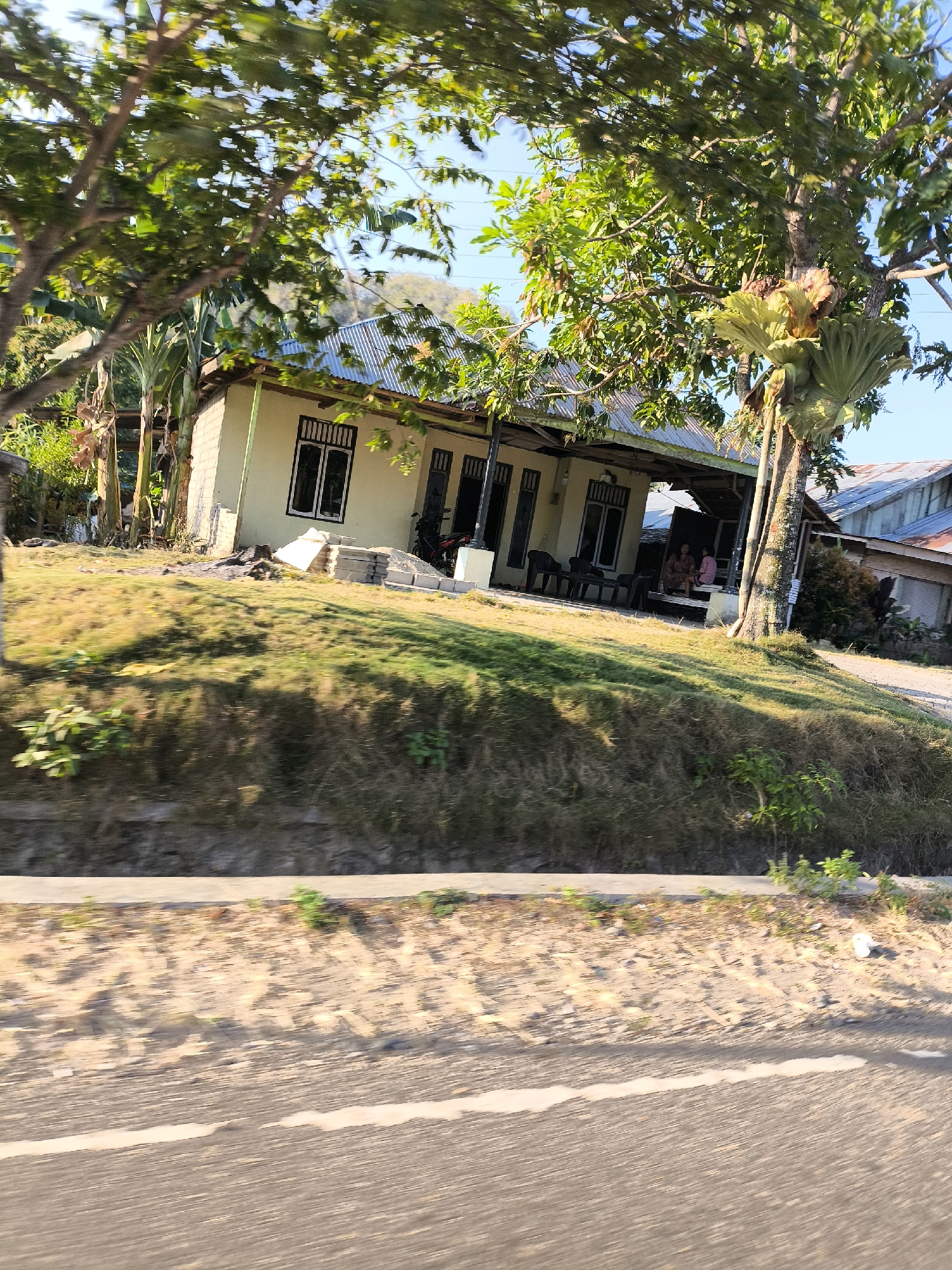 The image depicts a residential scene on a sunny day in Dulupi, Indonesia, as evidenced by the distinct architectural style of the house and the tropical vegetation. The primary subject is a single-story house with a light-colored exterior and a corrugated metal roof, typical of Indonesian homes. It features several windows with dark frames and a covered veranda area. In front of the house, there is a sloped lawn with patches of dry grass, indicating a warm climate. Several trees are visible, including a large one with an epiphytic plant growing on its trunk, adding to the tropical ambiance.
The house appears to be situated on a slight incline, with a road or pathway running parallel to the property in the foreground. The road has a concrete barrier along its edge and appears to be made of asphalt, with white markings indicating lanes. The ground beside the road is sandy and dry, with scattered patches of vegetation.
In terms of human presence, two individuals are seated on the veranda of the house, suggesting a relaxed, domestic setting. Their attire is not clearly discernible due to the image quality. There is also a motorcycle parked near the house, indicating common modes of transportation in the area. Construction materials, such as stacks of bricks or concrete blocks, are visible near the house, suggesting ongoing renovations or building work. The overall impression is that of a tranquil, semi-rural or suburban environment in Indonesia.