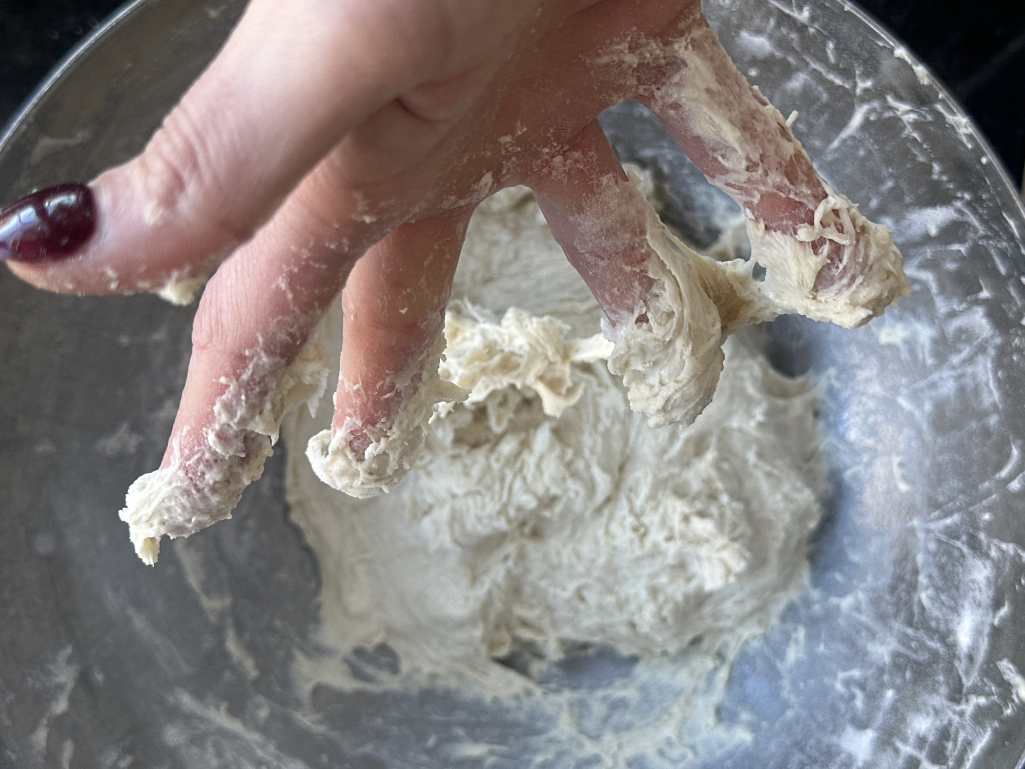 A human hand, covered in white dough, is seen above a metal bowl also containing a mass of white dough. The fingernail of the index finger is painted a dark burgundy color. The background is a dark, indistinct surface. The location is not discernible from the image.
