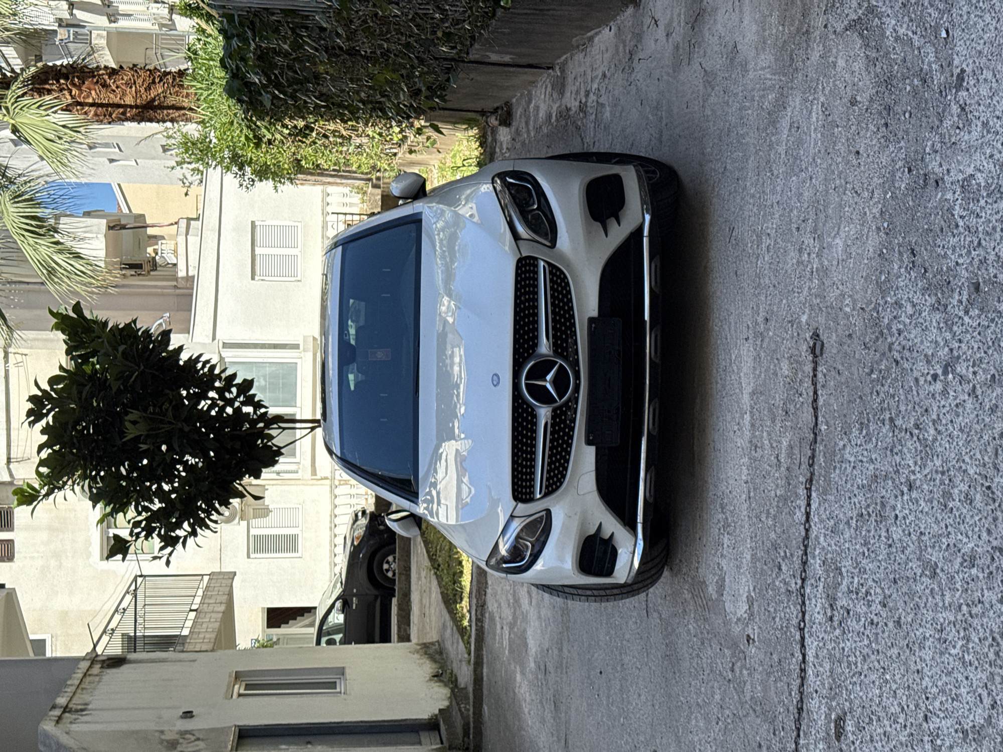 The image shows a white Mercedes-Benz GLC SUV parked on a concrete surface, likely a driveway or narrow street, in a residential area. The car faces the viewer head-on. Behind the car, there are several buildings, mostly apartment blocks, painted in light colors. A tree with dense foliage is visible to the left of the car, partially obscuring the building behind it. Another car is parked behind the white Mercedes. The setting suggests an urban environment, potentially in Budva, Montenegro, given the architectural style and vegetation visible. A small chain is running down the right-hand side of the concrete surface.