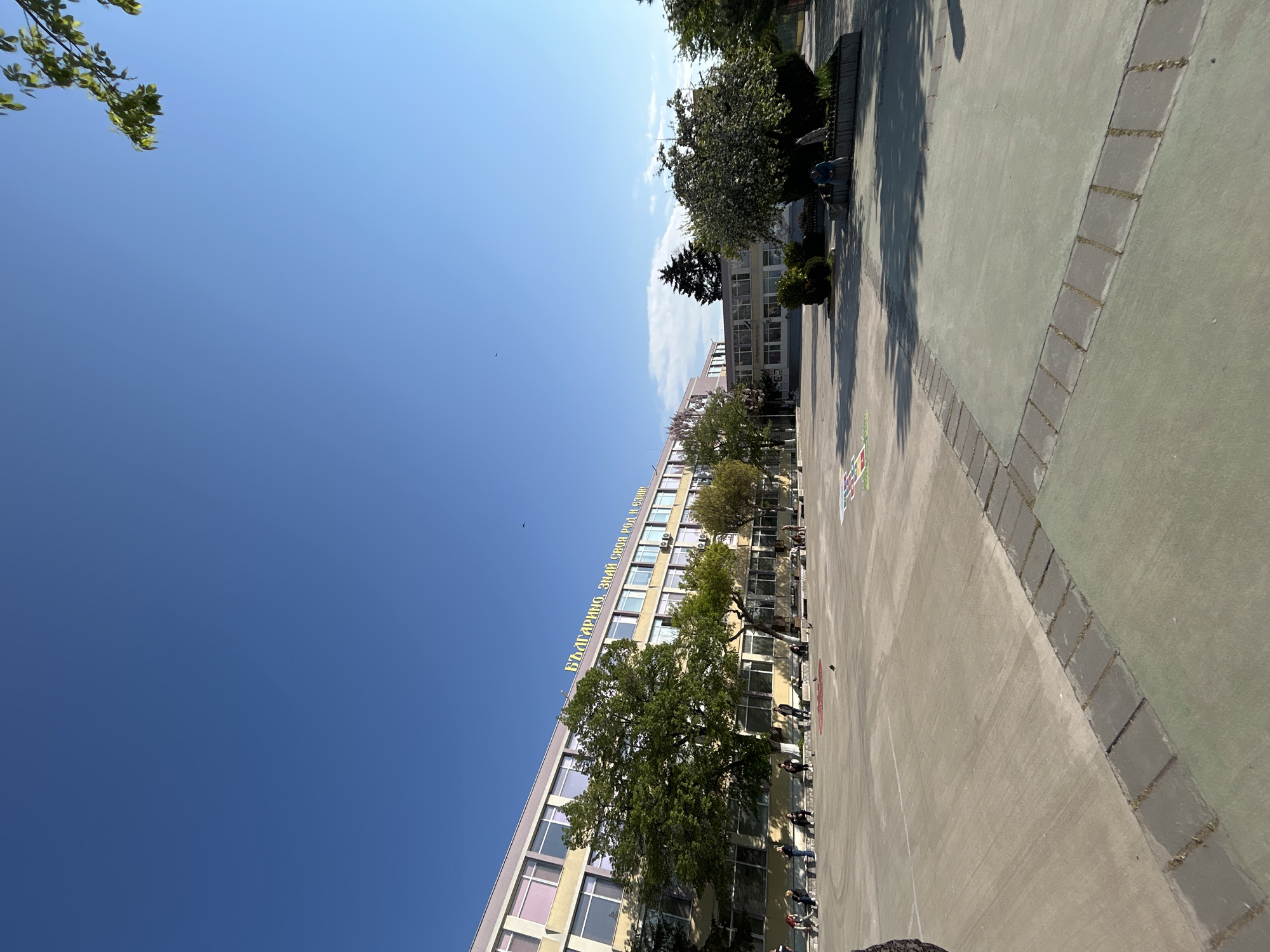 The image shows a tilted view of a building in Plovdiv, Bulgaria. The building, likely a school or public institution, has multiple floors with windows. A sign with text in Bulgarian is visible on the building facade. A large tree partially obscures the view of the building. People are visible standing along the edge of a paved area in front of the building. The paved area is marked with painted lines and patterns. The sky is blue with some wispy clouds.