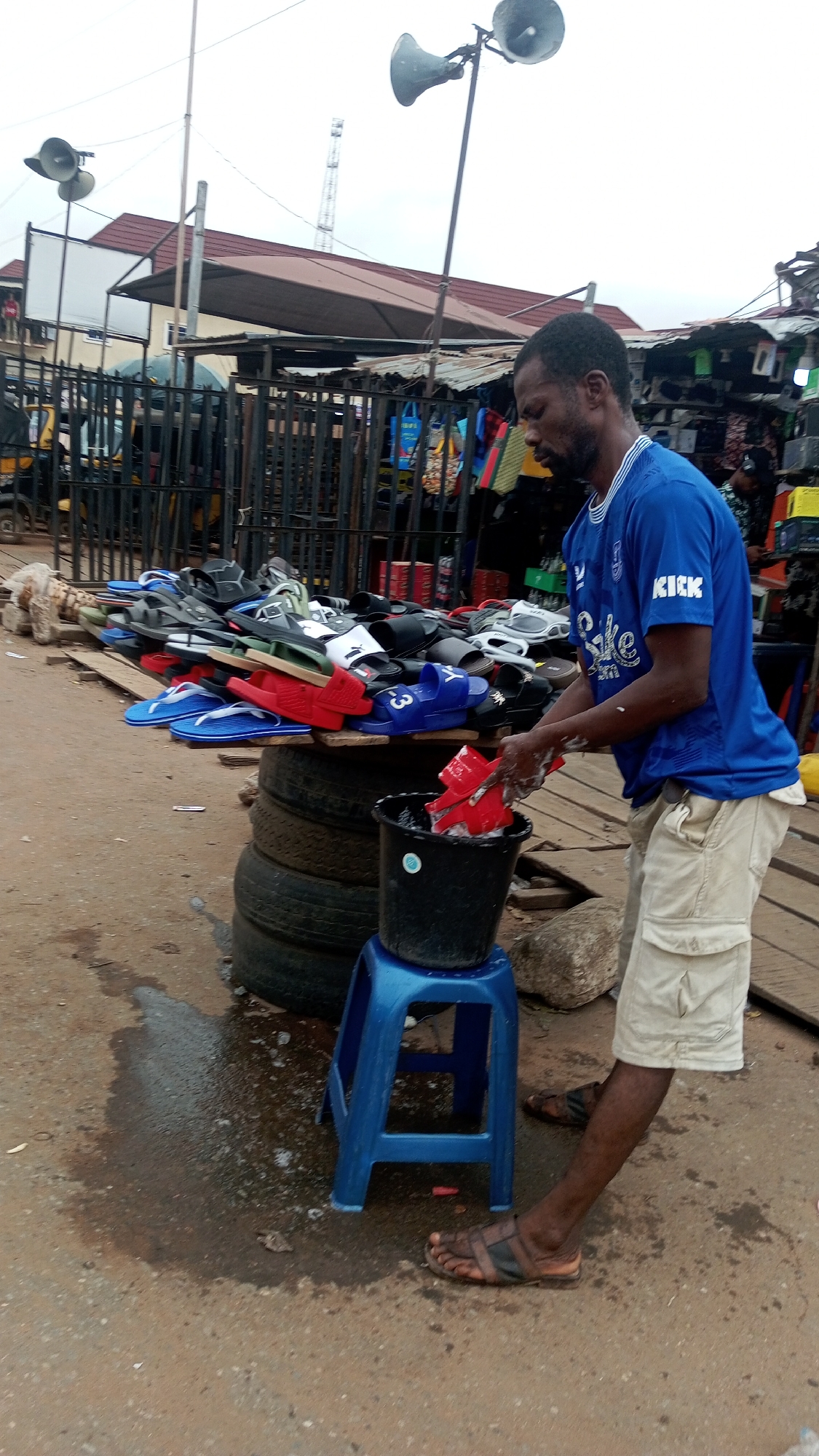 This image depicts an outdoor market scene in Onitsha, Nigeria, focusing on a street vendor preparing merchandise. The central figure is a dark-skinned man with short dark hair and a beard, dressed in a blue short-sleeved t-shirt with "KICK" visible on the sleeve, light-colored cargo shorts, and dark sandals. He is actively engaged in washing a red slide sandal in a black bucket, which is placed on a small blue plastic stool. His hands are wet and show traces of suds.
To the man's left, a makeshift stall displays a variety of slide sandals and flip-flops. The stand is constructed from wooden planks supported by stacked tires. The footwear, in colors such as red, blue (one pair has a "3" visible), black, white, and green, is neatly arranged for sale. The ground around the washing area is damp and appears to be dirt or worn pavement.
The background reveals a bustling market environment with dark metal fences, other market stalls displaying clothing and various goods, and partially visible vehicles. Overhead, multiple horn-shaped loudspeakers are mounted on tall poles, suggesting public announcements or music. In the distance, buildings with reddish-brown roofs and a communication tower are visible against an overcast sky, indicating it is daytime. The scene captures a moment of daily commerce in a vibrant market setting.