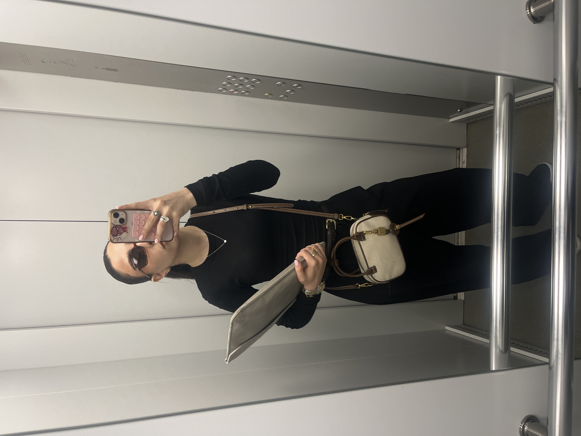 A woman is taking a selfie in a mirrored elevator. She is wearing sunglasses, a black long-sleeved top, and black pants. She has a small beige bag with a brown strap over her shoulder and is holding a grey laptop case. The elevator has white walls, a silver ceiling panel with round lights, and silver handrails. There is no indication of the location being in Yerevan, Armenia.