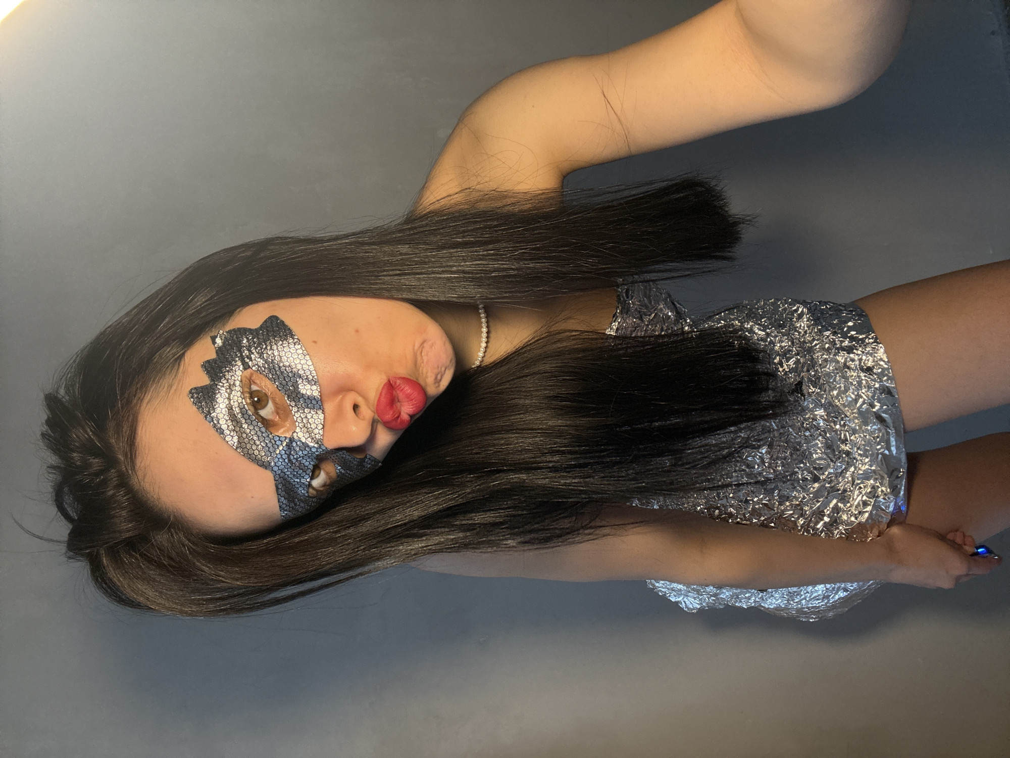 A woman is lying on a gray surface. She is wearing a silver sequin mask on her face. She has long, black hair, a pearl necklace, and red lipstick. She is wearing a silver dress, and her arms are above her head. The location in the city of Votkinsk, Russia, is unknown.