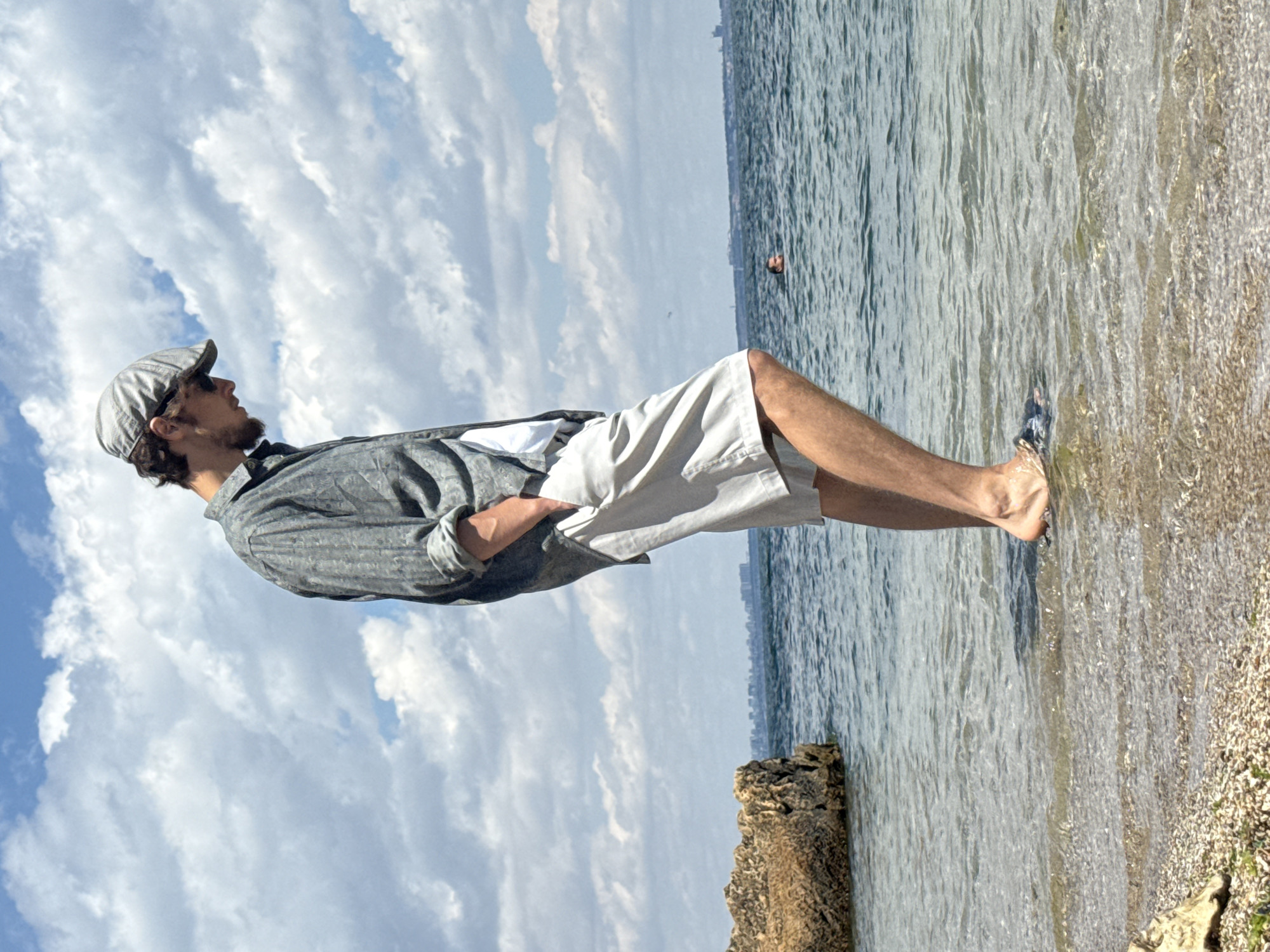 A man stands at the edge of the sea, his feet partially submerged in the shallow water. He is wearing a grey patterned flat cap, sunglasses, a grey short-sleeved button-down shirt over a white t-shirt, and light-colored shorts. His hands are in his pockets, and he is looking down. The water is clear and reflects the sky. In the background, a city skyline is visible across the sea, and a few clouds dot the blue sky. To the left of the man, a large rock juts out from the water. Further out in the sea, another person is swimming. The scene appears to be a beach or coastline within Odesa, Ukraine, on a bright, partly cloudy day.