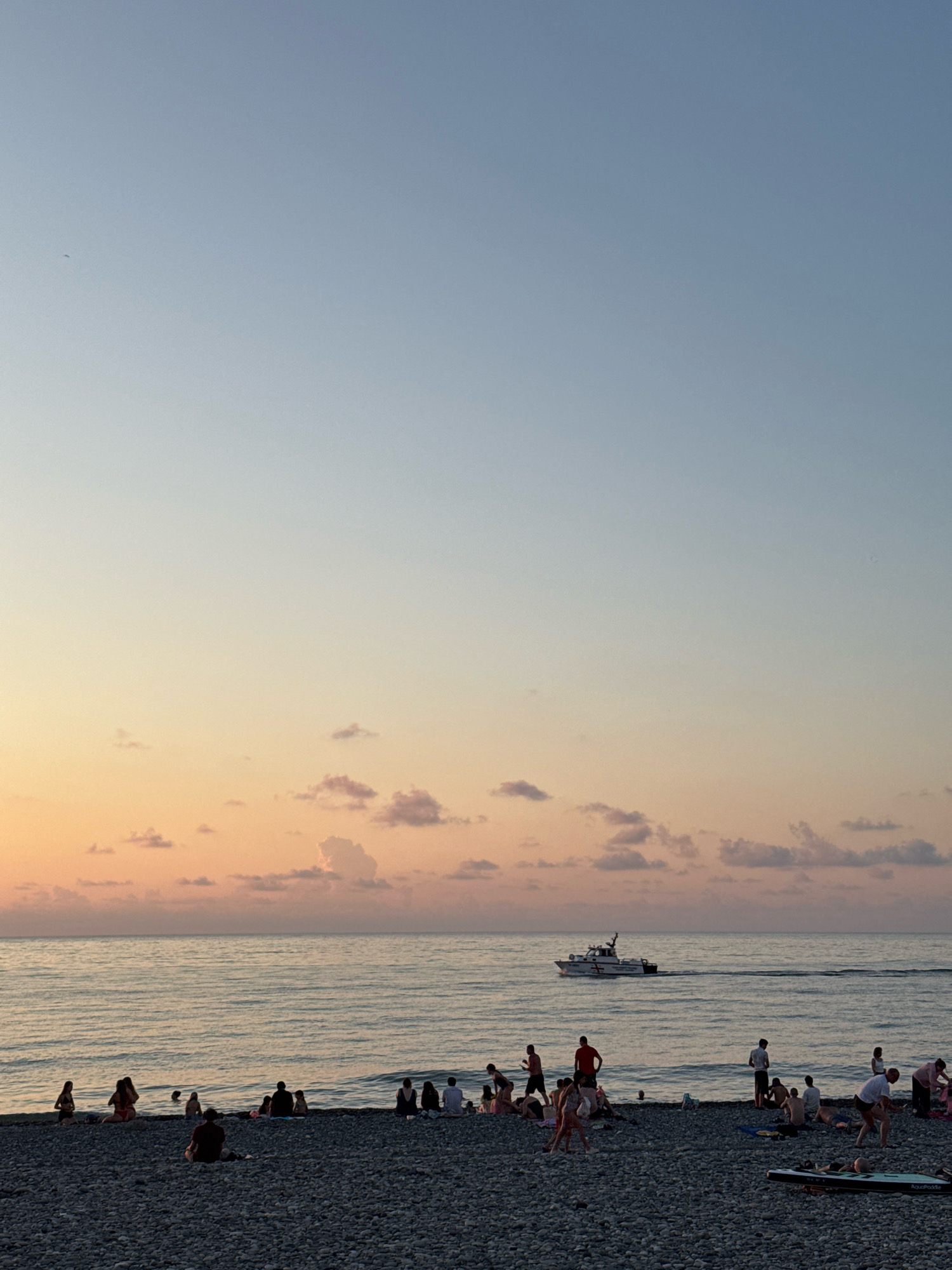 The image depicts a pebble beach scene in Batumi, Georgia, at dusk. The sky transitions from a pale blue at the top to soft hues of orange and pink near the horizon. Several small, scattered clouds are visible. The sea is calm with gentle ripples. A white boat with red markings is navigating the water. On the beach, numerous people are casually gathered, some sitting, others standing or walking. A few individuals are partially in the shallow water. The beach is covered in dark grey pebbles. The overall atmosphere is serene, characteristic of an evening at the seaside.