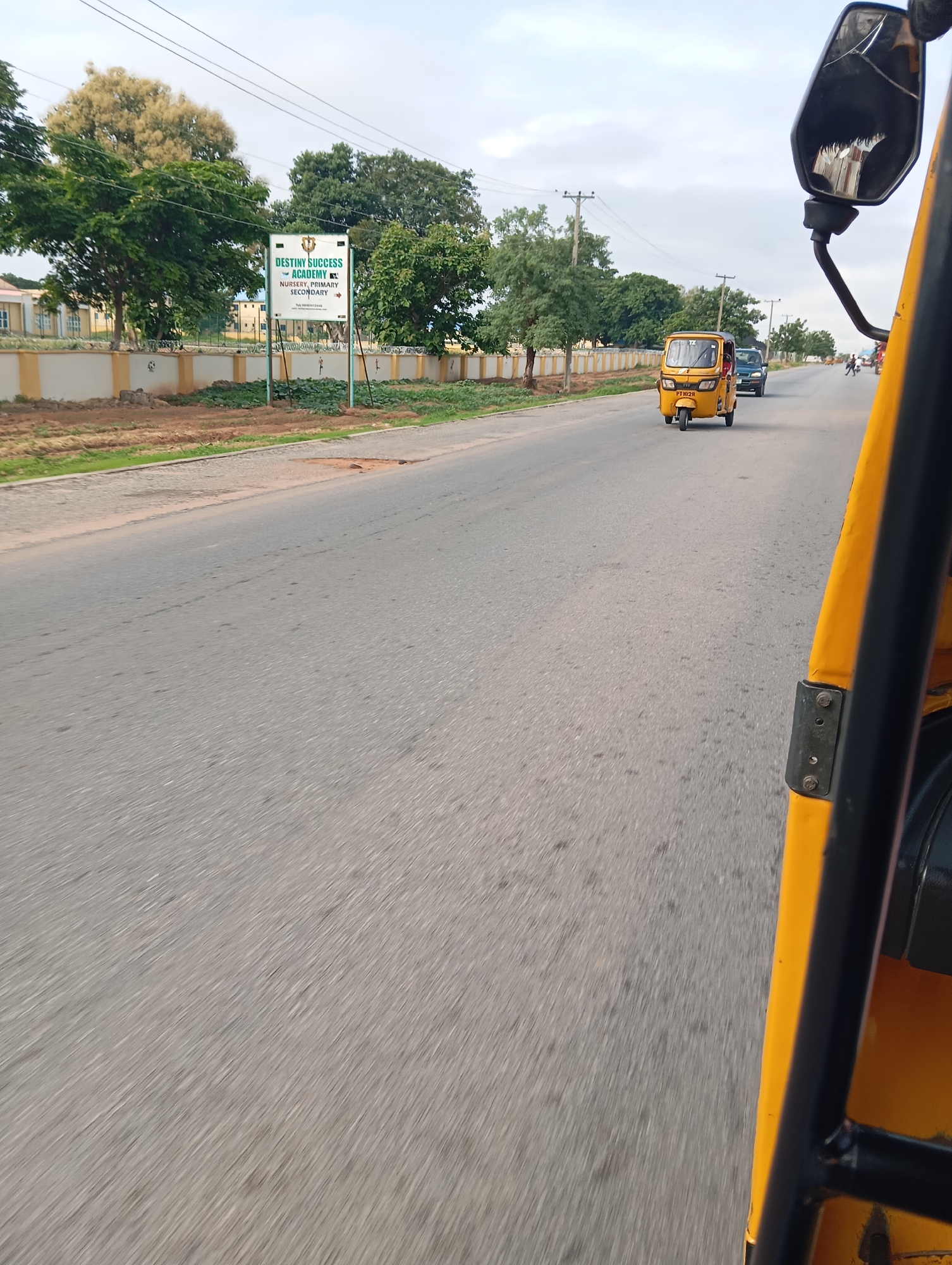 The image depicts a street scene in Jalingo, Nigeria. In the foreground, the right side of a yellow auto-rickshaw is visible, with its black metal frame and a portion of the side mirror. The view is from within the auto-rickshaw, looking forward and to the left.
On the left side of the road, a school sign for "DESTINY SUCCESS ACADEMY" is prominently displayed. The sign indicates that the academy offers nursery, primary, and secondary education. Behind the sign, a light-colored building with a yellow stripe runs along its base, likely part of the school. Lush green trees are situated between the road and the school building.
The road itself is paved with asphalt and appears to be a two-lane highway. Several vehicles are visible on the road, including a yellow auto-rickshaw ahead of the camera vehicle, and a dark-colored car further down the road. The sky is overcast with clouds. The environment suggests a daytime scene, possibly in the late afternoon or early morning, given the lighting conditions. The presence of electric poles and overhead wires indicates that this is a developed area within the city. The overall impression is of a typical urban or peri-urban street in Nigeria.