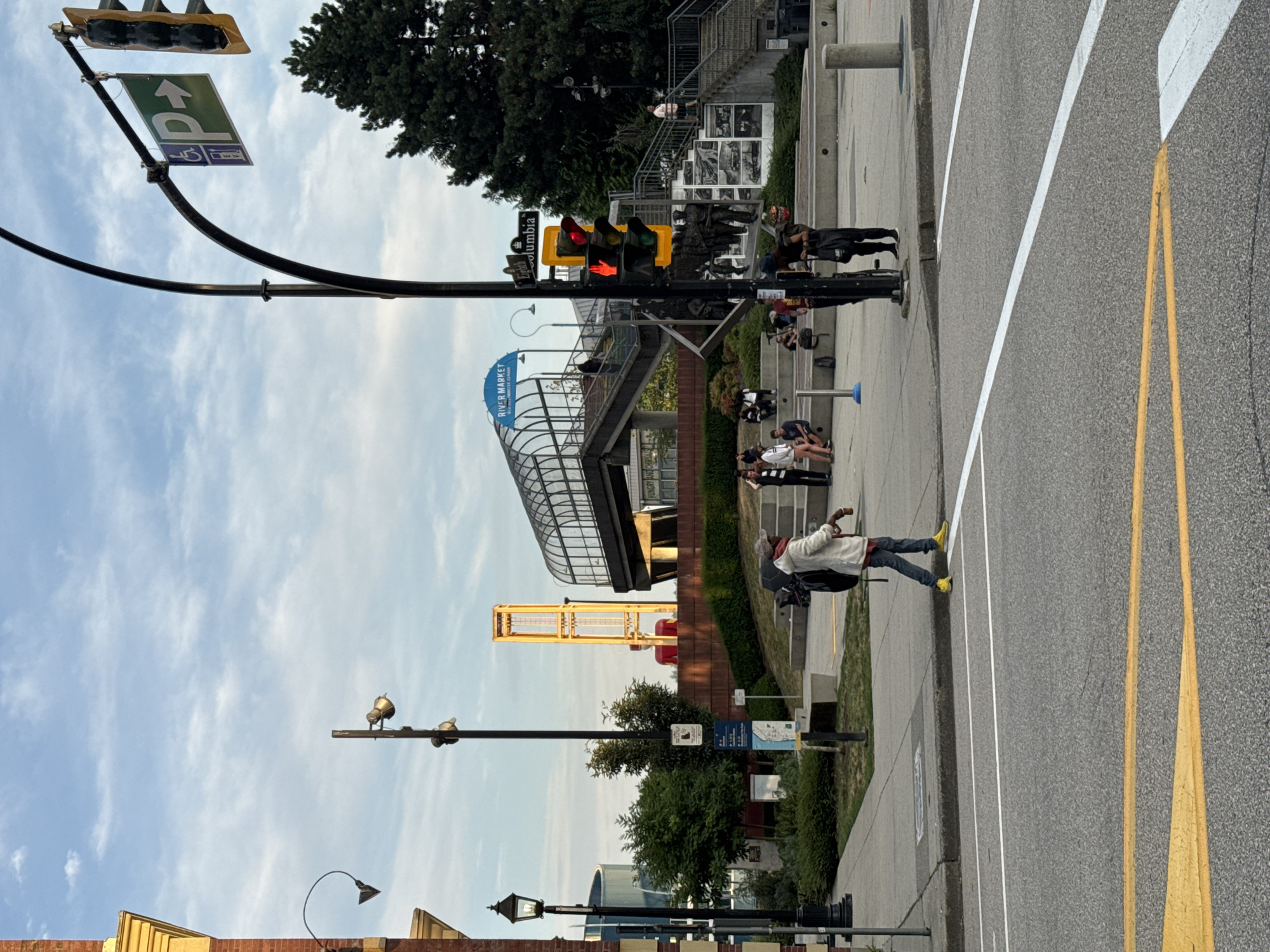 The image depicts a street scene in the city of New Westminster, Canada, likely near the River Market. In the foreground, a person wearing a white coat, dark pants, and yellow shoes walks across a street intersection. The street has white and yellow lane markings.
In the background, a traffic light is visible, displaying a red hand signal for pedestrians. Above the traffic light, a sign indicates "Eighth Columbia". Further in the background, there is an elevated walkway with a glass enclosure, leading to a structure labeled "RIVER MARKET". Several people are seated on tiered steps in front of the walkway. A tall yellow tower structure is also visible in the distance, along with trees and a cloudy sky. A black lamppost with multiple lights stands near the center of the image. Signs on poles near the lamppost indicate directions and information, including an accessibility symbol and a parking symbol with an EV charging icon.
