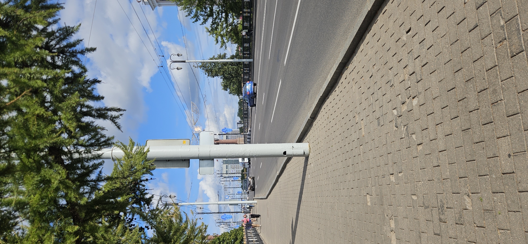 The image shows a street scene on a bridge in Vitsebsk, Belarus. In the foreground, a paved sidewalk runs along the left side of a multi-lane road. A large, grey metal pole, possibly a lamppost or traffic signal pole, stands on the sidewalk, partially obscuring the view of the road. Green foliage from a tree hangs over the scene from the top left.
On the sidewalk, several people are walking. The road has multiple lanes and is carrying traffic. A dark-colored car is in the middle lane, and a blue car is in the right lane, further down the road. On the right side of the road, a decorative metal railing lines the edge of the bridge. In the background, buildings and more lampposts are visible. The sky is blue with scattered white clouds. The overall impression is of a sunny day on a busy urban bridge.
