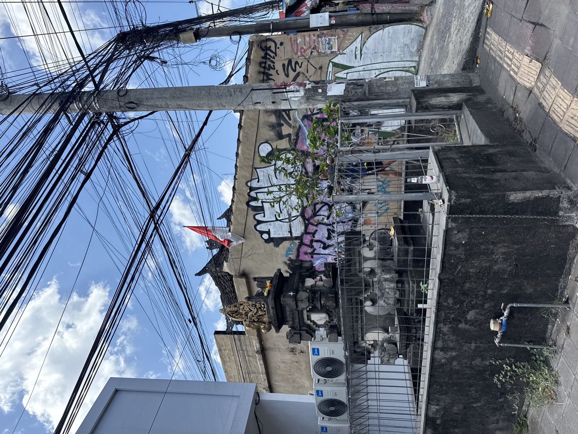 In North Kuta, Indonesia, a street scene unfolds under a bright blue sky dotted with white clouds. A complex network of black electrical wires crisscrosses the upper portion of the frame, suspended from a tall concrete utility pole on the right. To the left, a white building with a smooth facade is visible. In the center of the image, a traditional Balinese shrine, constructed from dark stone, stands prominently. It is adorned with intricate carvings and topped with a golden statuette. A small offering of flowers is placed on the shrine. Behind the shrine, a building with a tiled roof is covered in graffiti. A red and white Indonesian flag is attached to the wall. In front of the shrine, a metal fence encloses the area. Two white air conditioning units are mounted on the wall to the left of the shrine. The street in the foreground is paved with grey concrete tiles, and a textured tactile paving strip runs along the edge. Green vegetation is growing in patches along the side of the path and against the wall of the shrine.