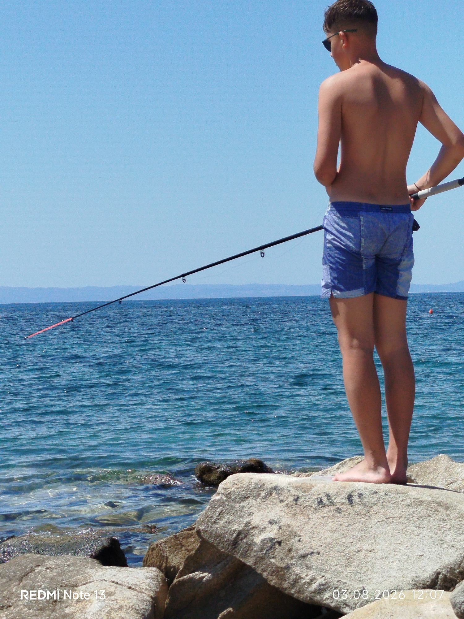 A young male, viewed from behind and from the waist down, stands on large, grey rocks next to the sea. He wears blue denim shorts and sunglasses. He is holding a fishing rod, which extends over the water. The sea is a vibrant blue, with small waves. In the distance, hazy blue mountains can be seen against a clear blue sky. The timestamp "03.08.2026 12:07" and "REDMI Note 13" are superimposed on the lower right and left corners of the image, respectively. The location is Sithonia Municipal Unit, Greece.