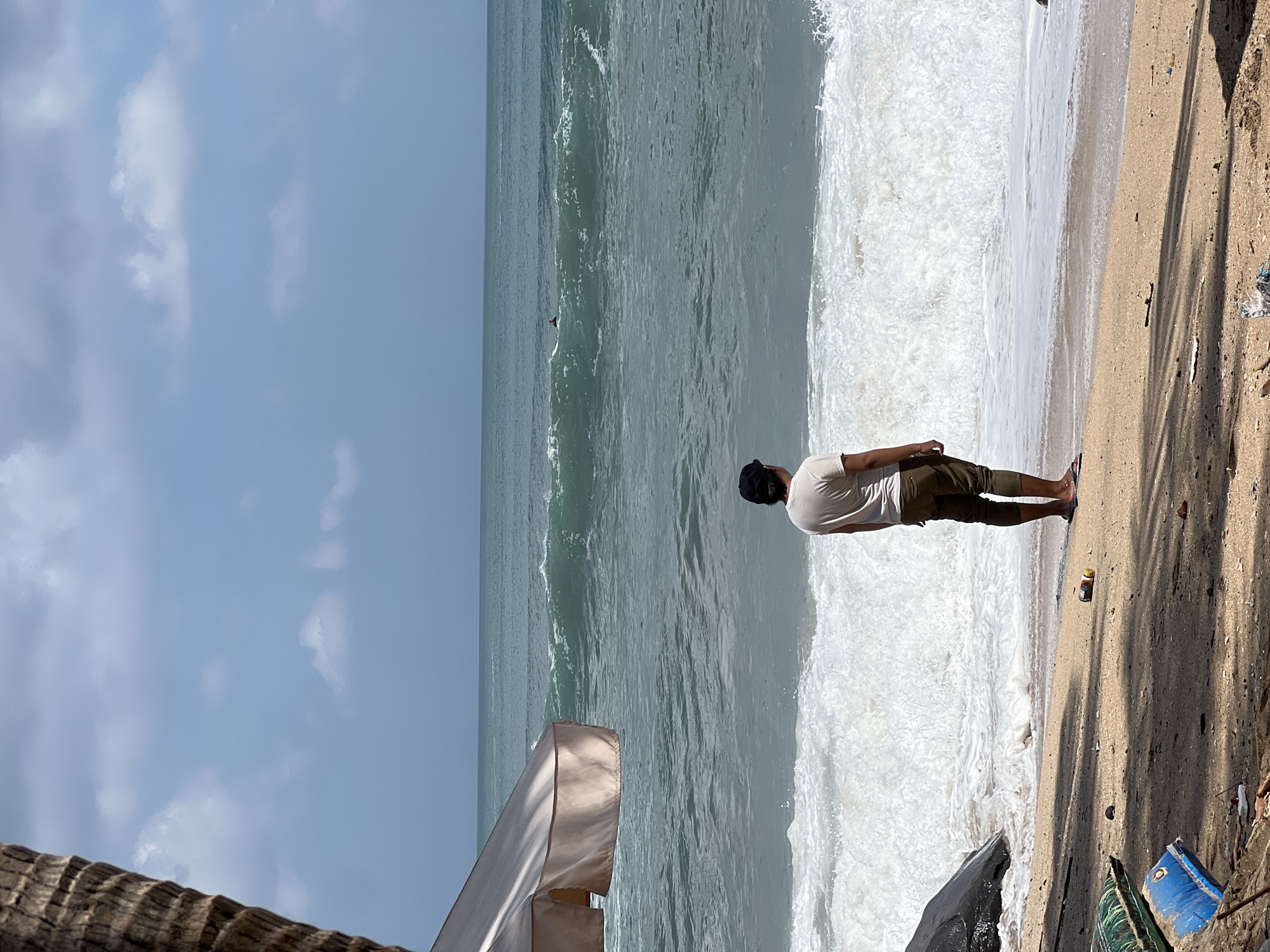 A man stands on a sandy beach in Kuta, Indonesia, facing the ocean. He is wearing a light-colored t-shirt and shorts, with a black baseball cap. The waves are crashing on the shore, and there is a surfer in the distance. A large palm tree trunk is visible on the left side of the frame, and a portion of a beach umbrella is also present. Debris, including plastic bottles and wrappers, is scattered on the sand. The sky is partly cloudy.