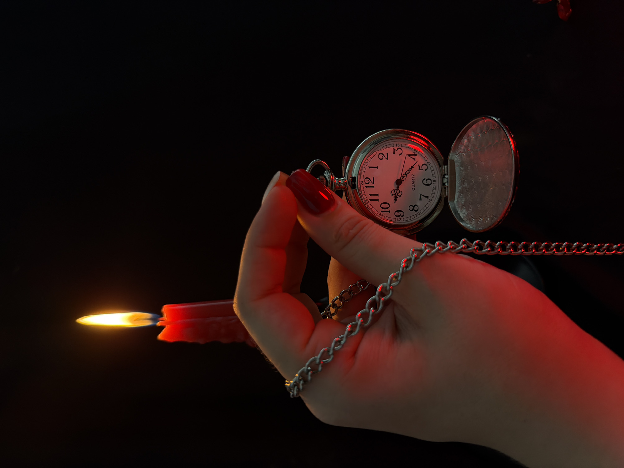 A close-up shot shows a hand holding a pocket watch. The watch is silver with a white face and Roman numerals. The time is approximately 9:45. The watch is attached to a silver chain. The hand has red nail polish. In the background, a red candle is lit. The scene is dark with a red hue cast by the candle. The image appears to be taken in an indoor setting.