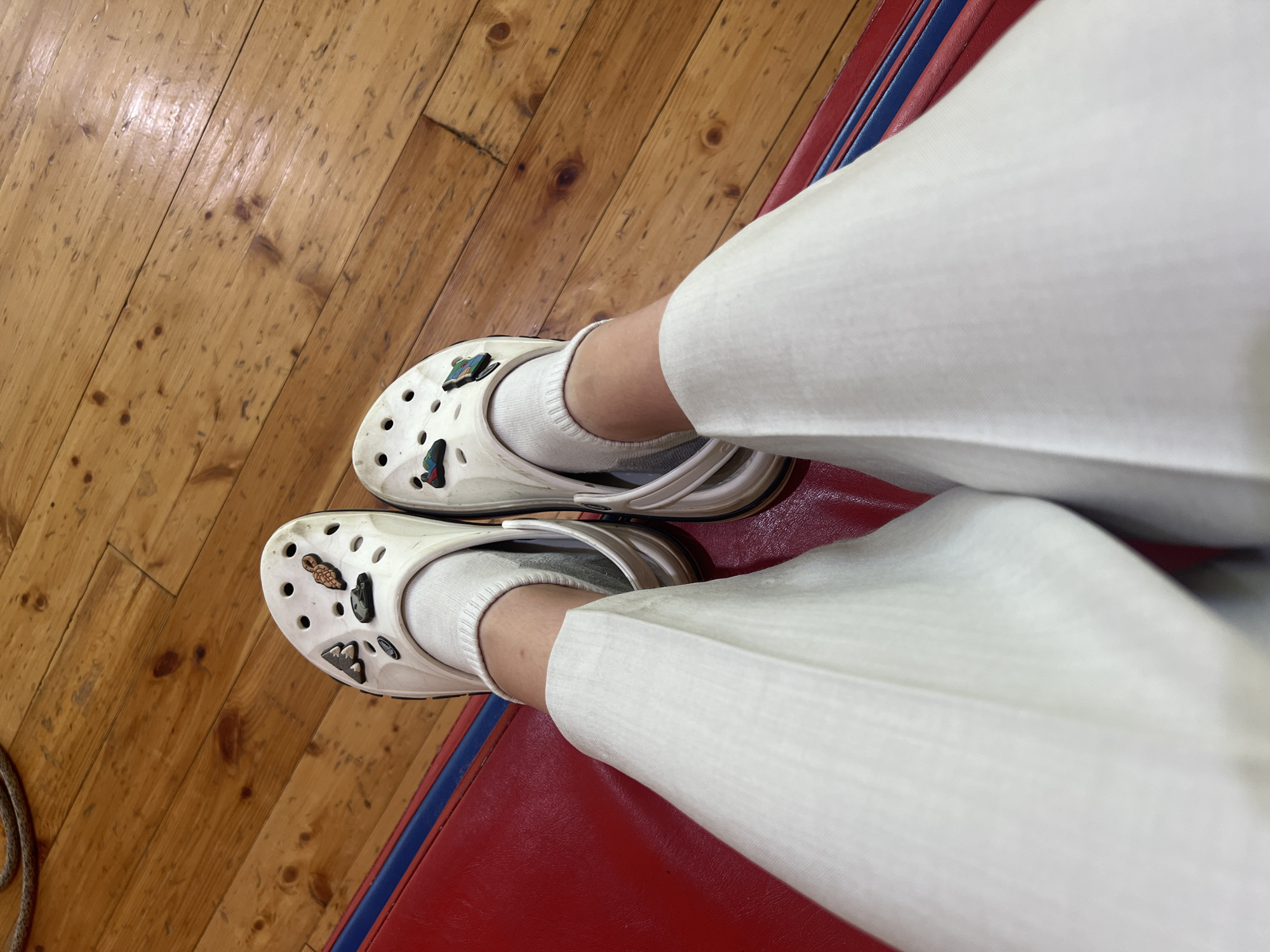 The image shows a first-person perspective of a person's legs and feet. The person is wearing white Crocs with various charms, white socks, and white pants. The person is standing on a wooden floor, with a red mat with blue stripes visible on the right side of the image. It is impossible to determine if this is in Chaikovsky, Russia from the image.